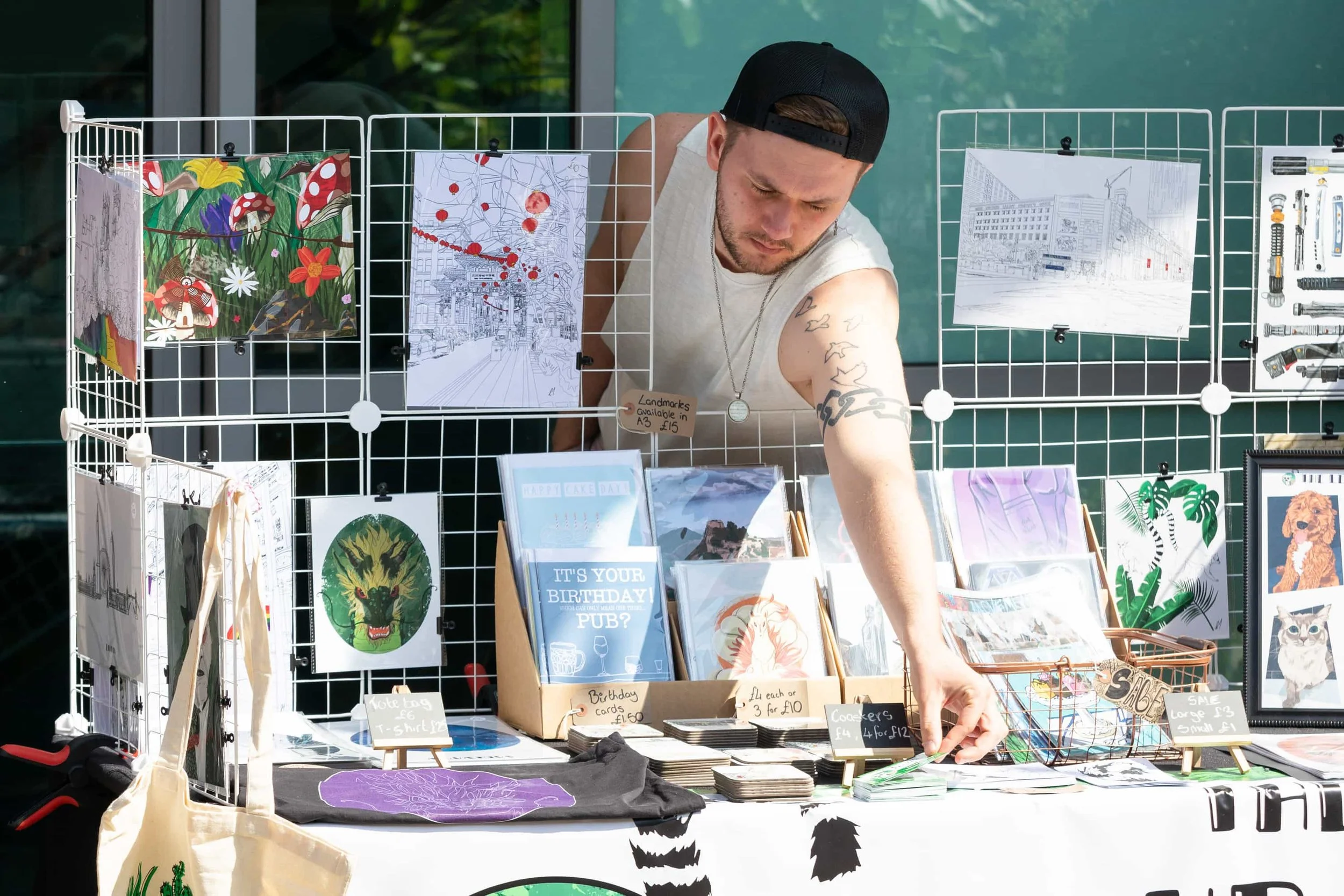 A man browsing through artwork and greeting cards at an outdoor art market stall with various illustrations and framed pictures on display.