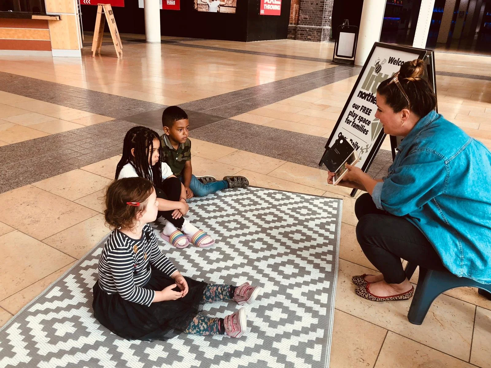 A woman sitting on a small stool in a shopping mall is talking to three children sitting on a patterned rug. The woman is holding a tablet and facing the children. The background shows mall storefronts and a signboard.