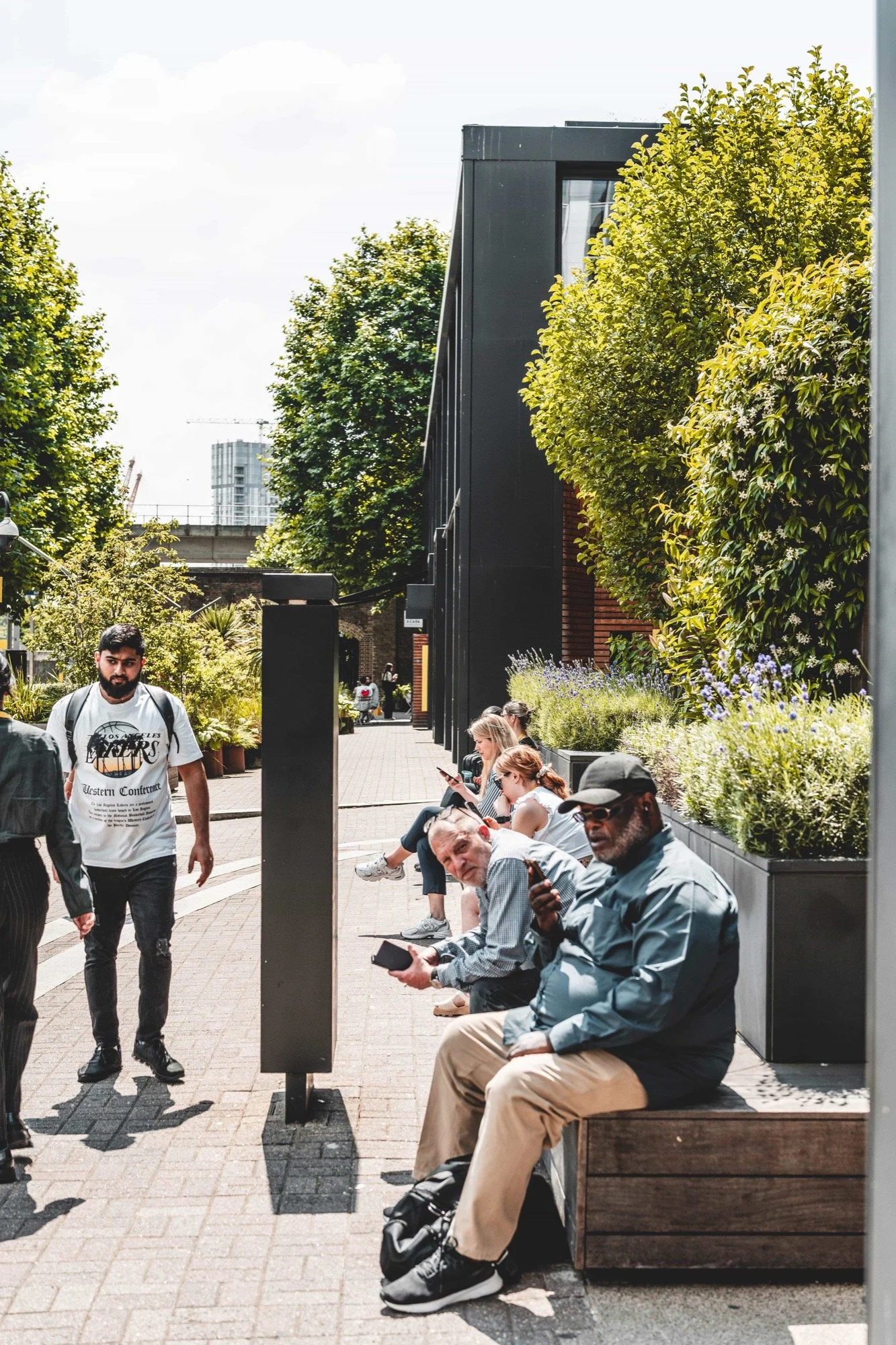 People sitting and standing on a sidewalk in an urban area with greenery and modern buildings in the background.