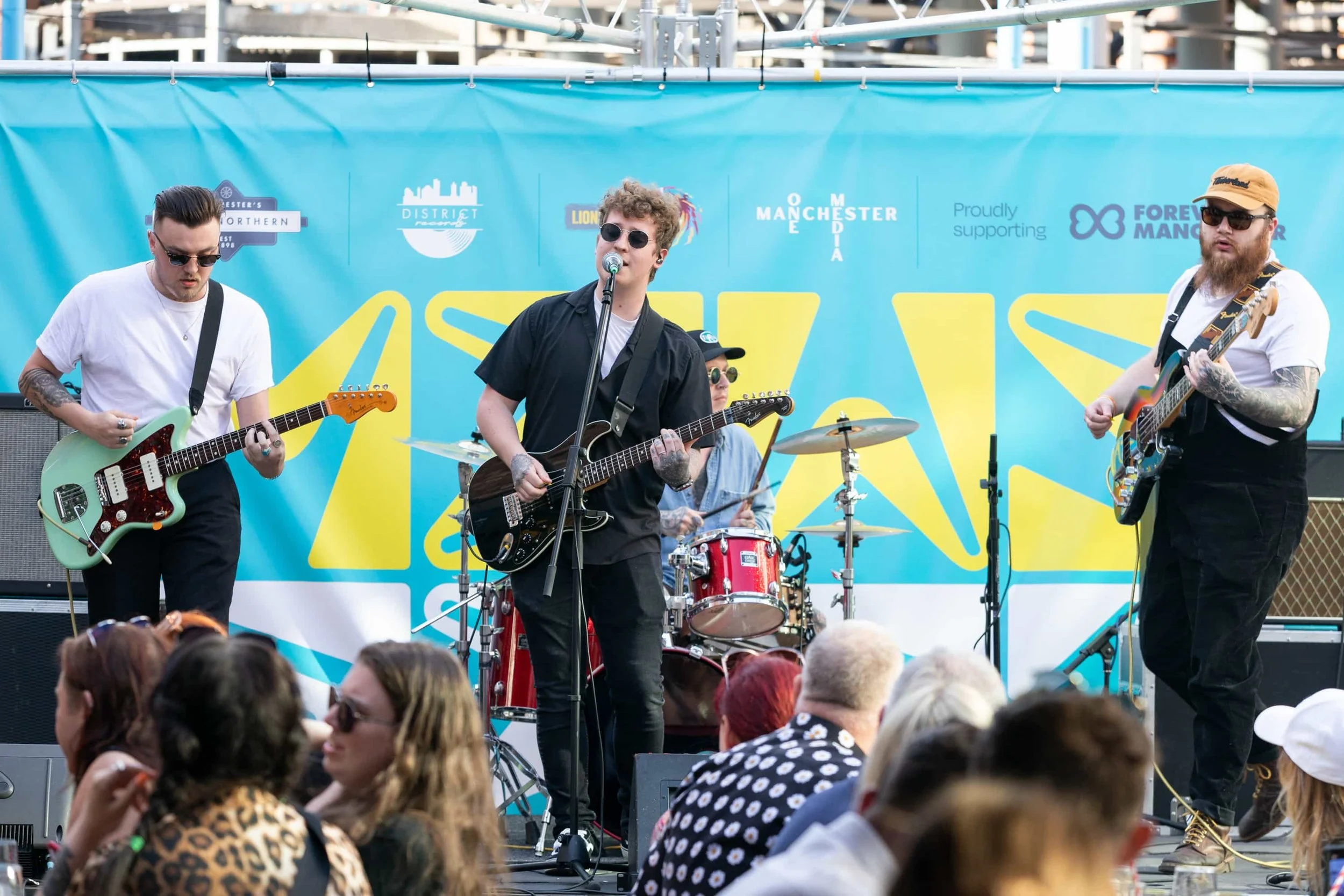 A band of four musicians performing on an outdoor stage during daytime at a music festival. Two guitarists and a drummer are visible, with an audience watching in front of the stage. The background features a bright blue banner with event logos and sponsors.