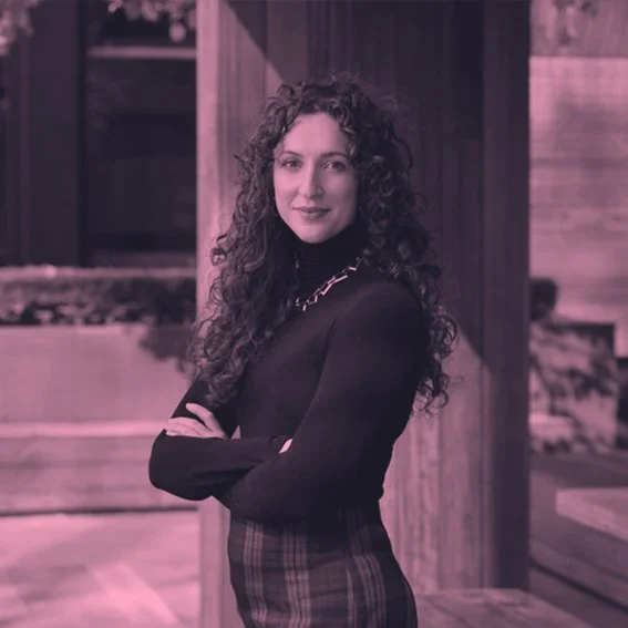 A woman with long curly hair standing indoors in front of wooden panels, wearing a black turtleneck and a plaid skirt, with her arms crossed.