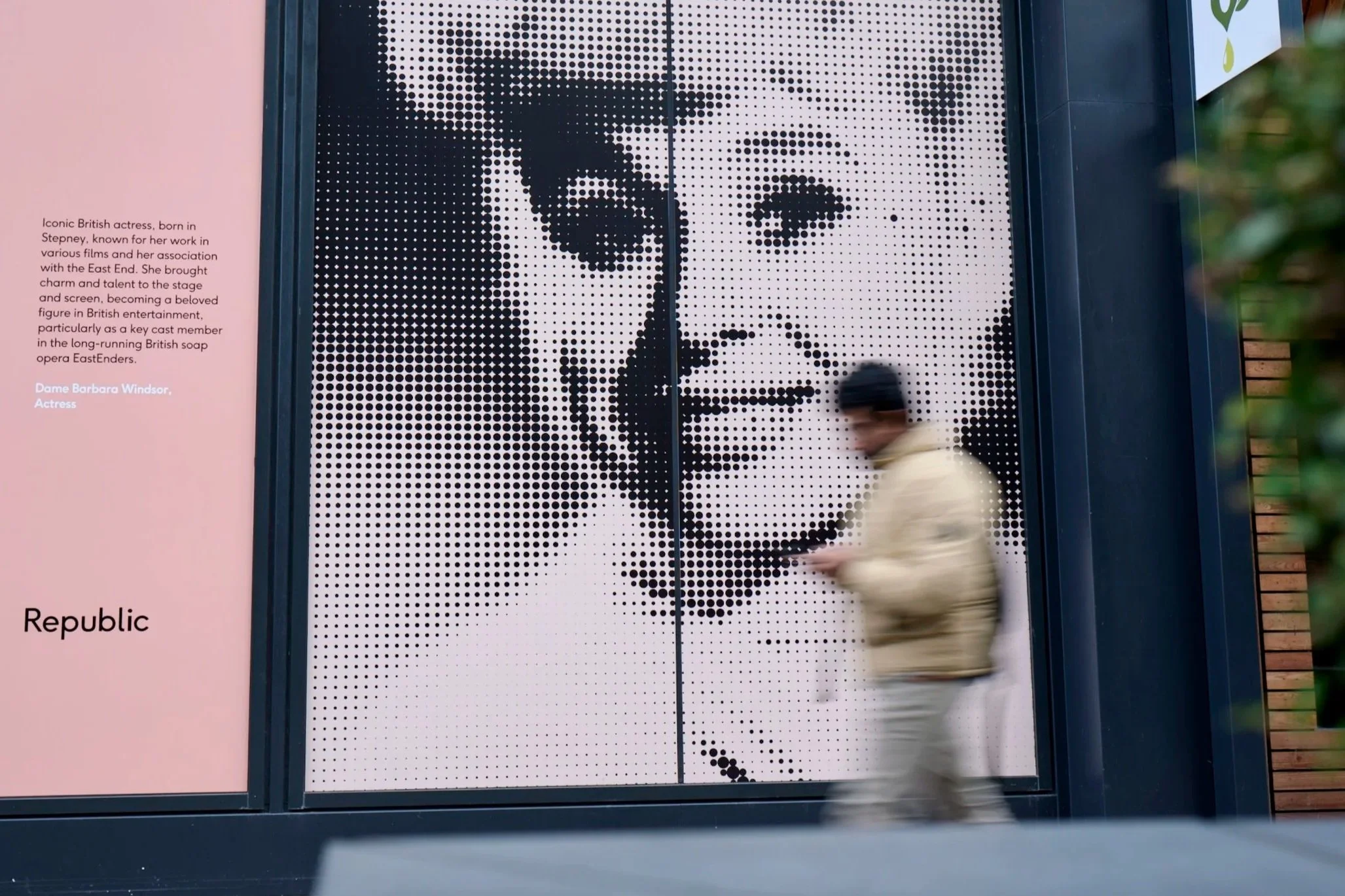 A large black and white pixelated portrait of a woman on an outdoor display, with a person in a beige jacket walking past in the foreground.