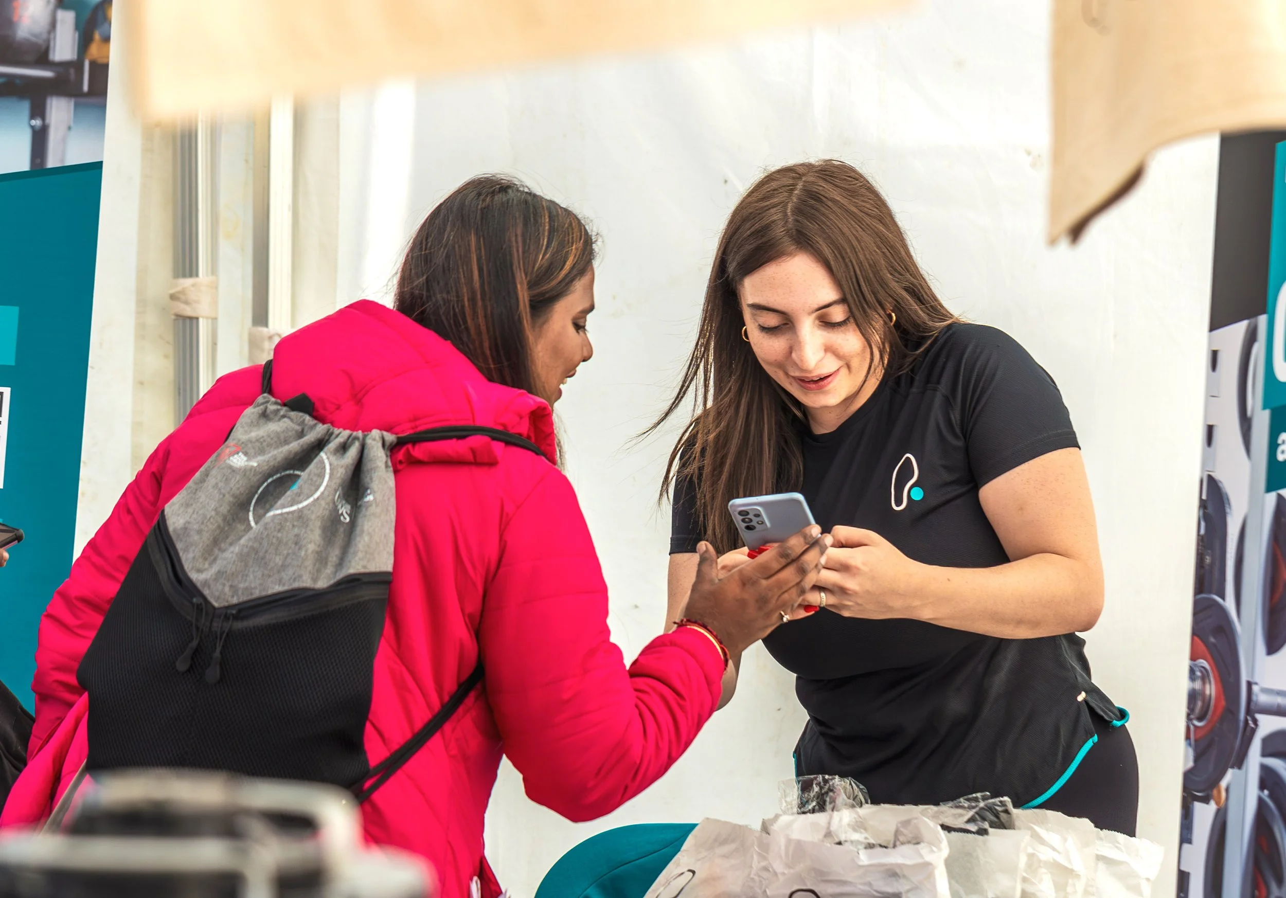 A woman in a black shirt shows a smartphone to another woman in a pink jacket, both looking at the phone. They are standing at a table with some paper and objects, possibly inside a store or booth.