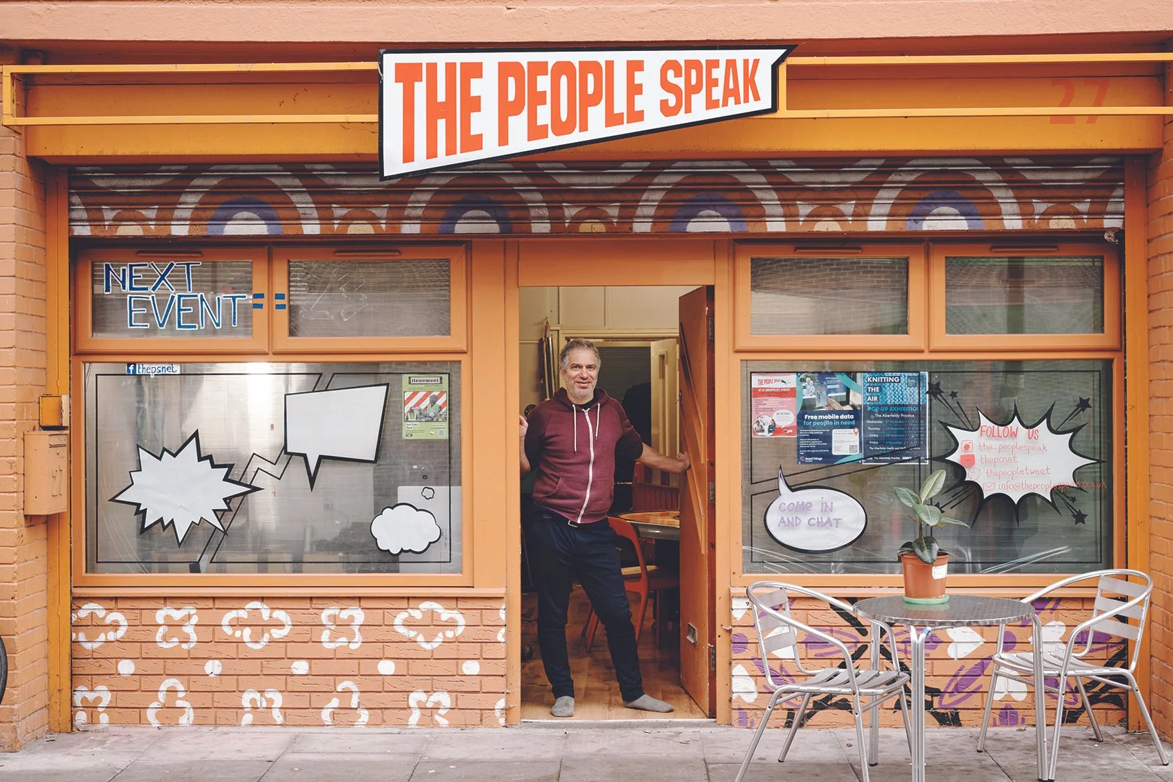 A man standing at the entrance of a building painted in orange with colorful graffiti and comic-style stickers on the windows. The signage reads 'The People Speak' and 'Next Event.' There are outdoor tables and chairs with a potted plant in front.