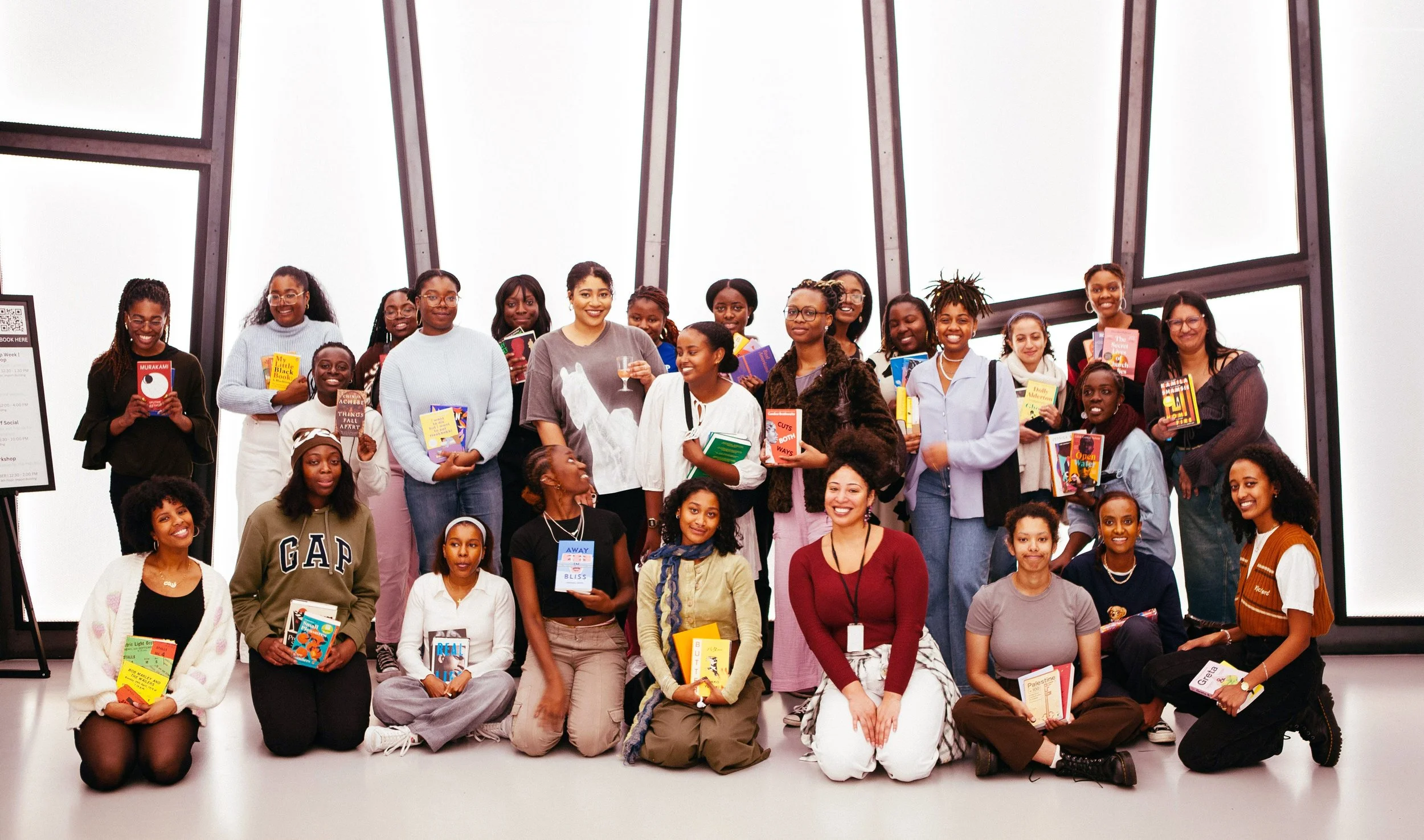 Group of women and girls posing with books in a bright indoor space with large windows.