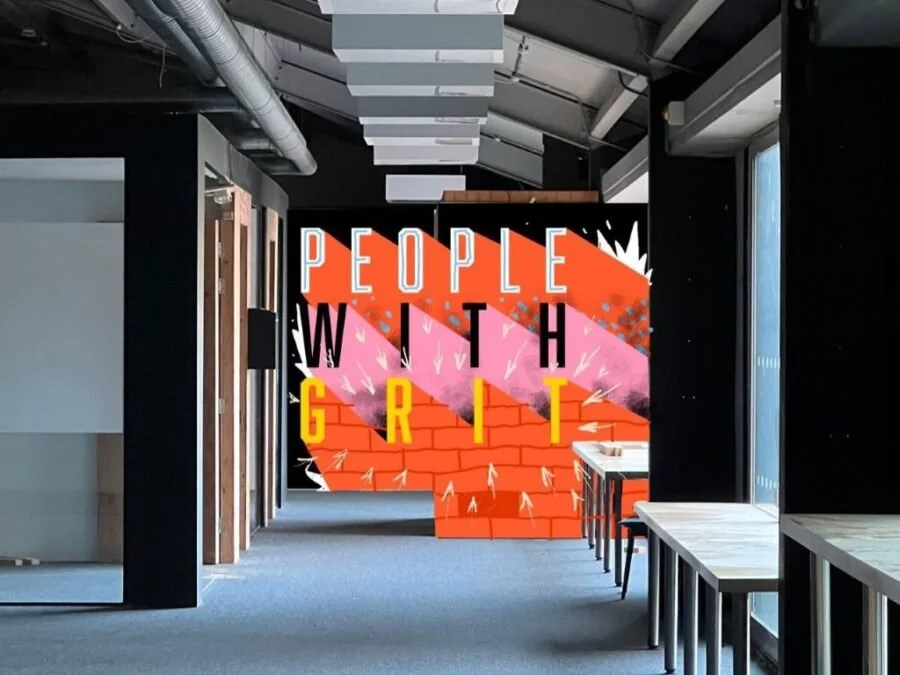 Interior of a modern office or coworking space with a colorful wall mural that reads 'People with Grit,' featuring graffiti style art, exposed ductwork, and windows with natural light.