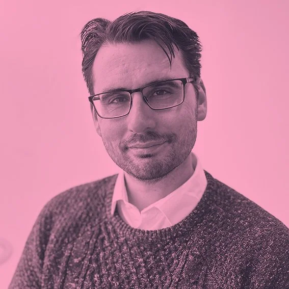 A man with dark hair, glasses, and a beard, wearing a white collared shirt and a dark sweater, against a pink background.