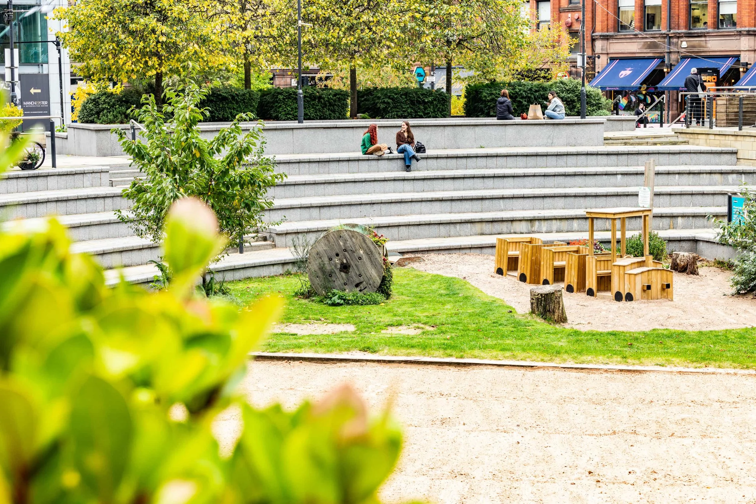 A small urban park with a playground featuring wooden train-shaped structures, a grassy area, trees, and stone steps with people sitting and walking nearby.