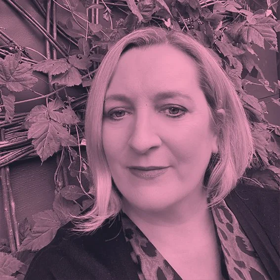 A woman with blonde hair taking a selfie in front of leafy vines and a woven fence.