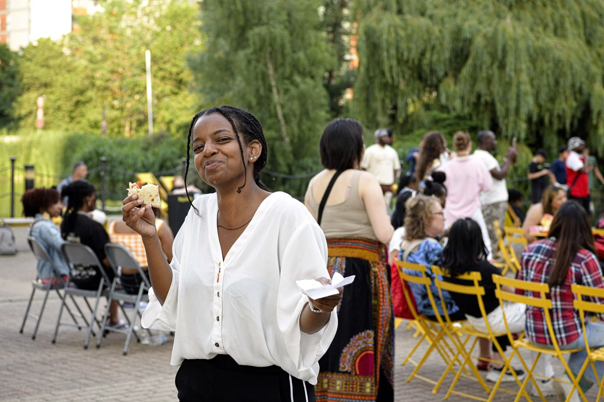 A woman in a white blouse holding a slice of cake and a napkin, smiling at a gathering in a park with trees and several people sitting on yellow chairs and standing in the background.