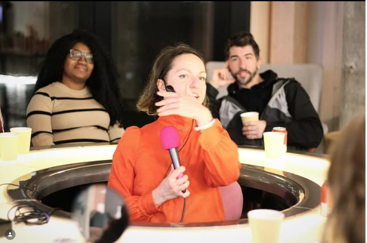 Three people sitting around a circular table. A woman in an orange jacket is holding a pink microphone and covering her mouth, smiling. A woman with glasses and a striped shirt, and a man with a beard and black jacket holding a yellow cup, are in the background.