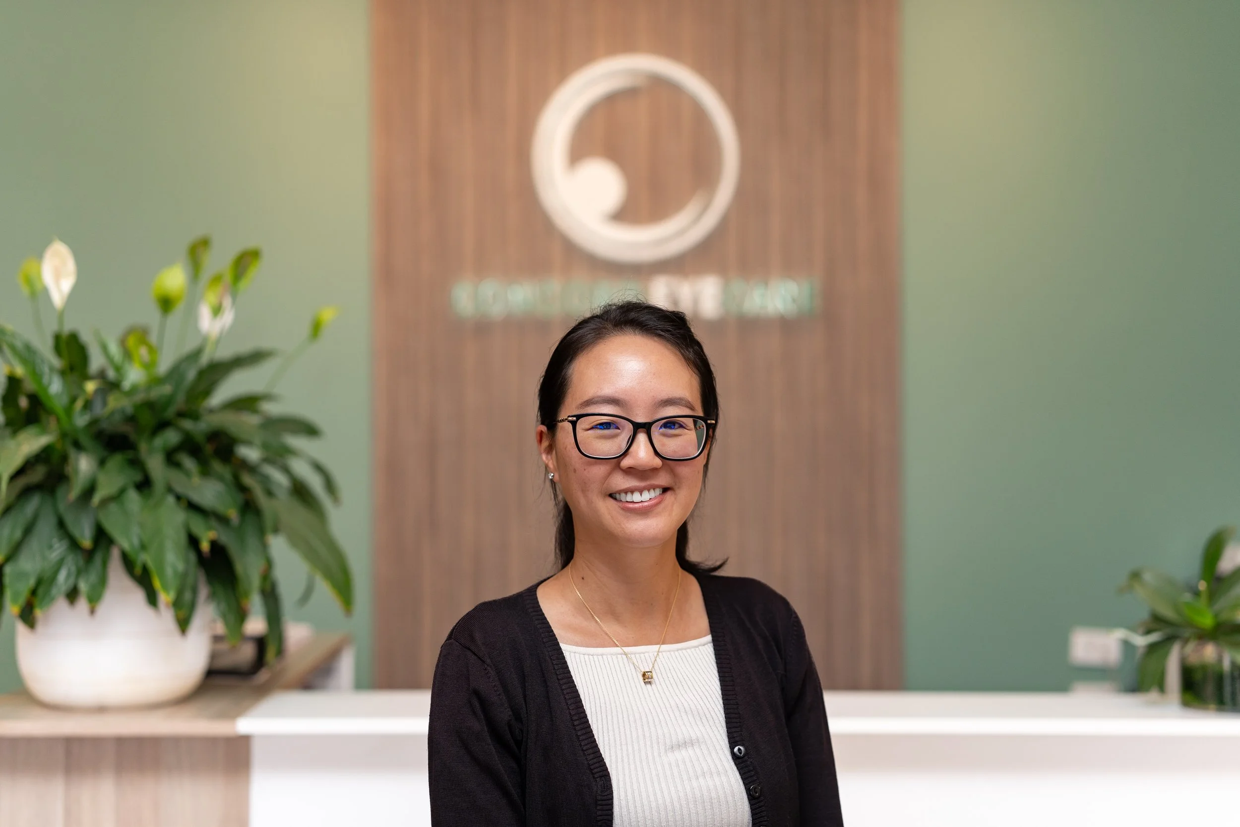 Optometrist Vivian Li with glasses smiling at the camera in an office lobby with green and brown walls, a flower plant on the left, and a wooden sign in the background.