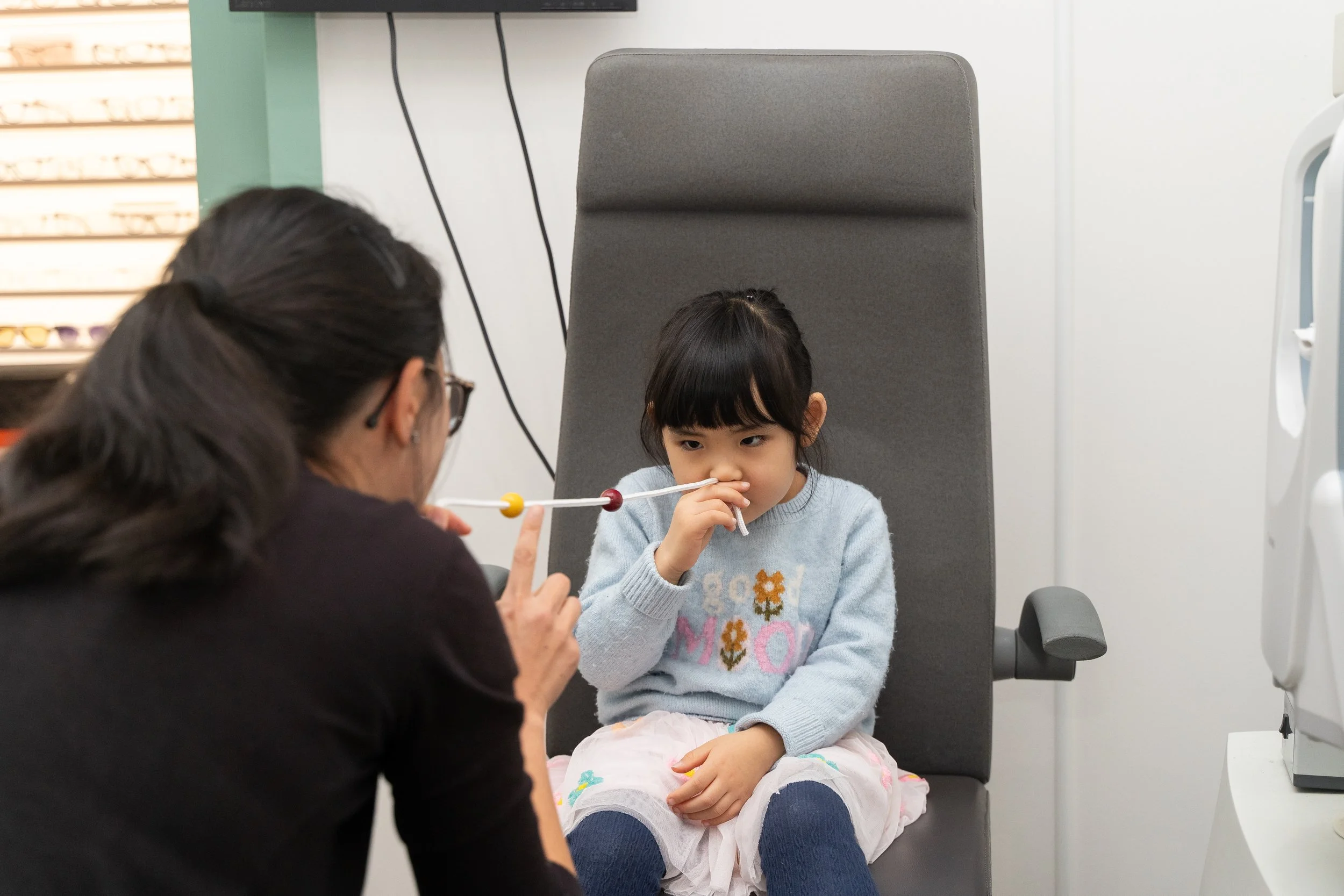 A young girl sits in an eye doctor’s chair, holding her nose, during an eye examination. An optometrist or ophthalmologist, partially visible with glasses, interacts with her using a red and yellow instrument.