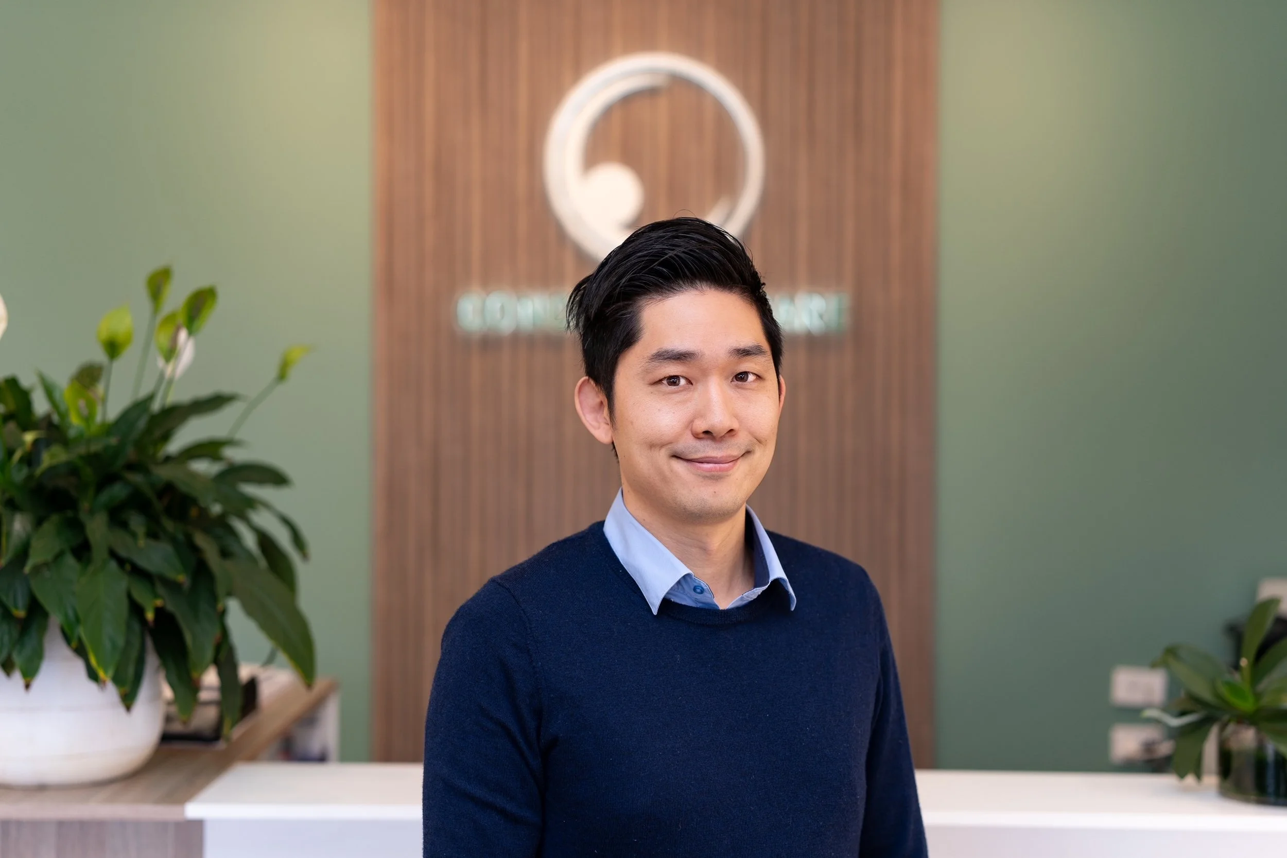 Optometrist Mark Joung with black hair in a blue sweater and collared shirt stands in an office lobby with a wooden wall, potted plants, and a blurred logo and text in the background.