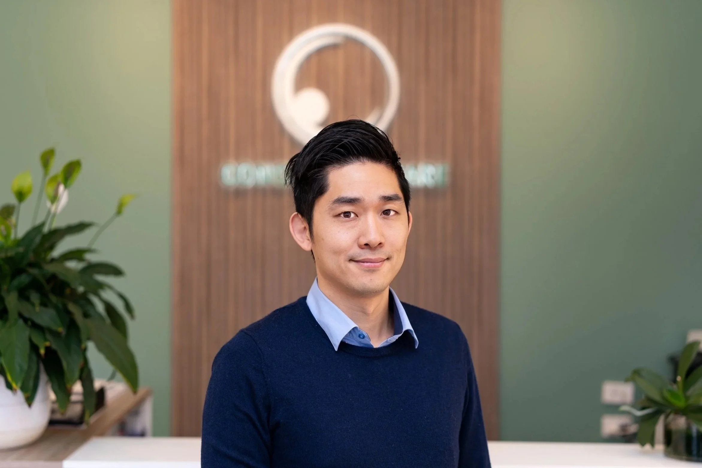 Optometrist Mark Joung with black hair in a blue sweater and collared shirt stands in an office lobby with a wooden wall, potted plants, and a blurred logo and text in the background.