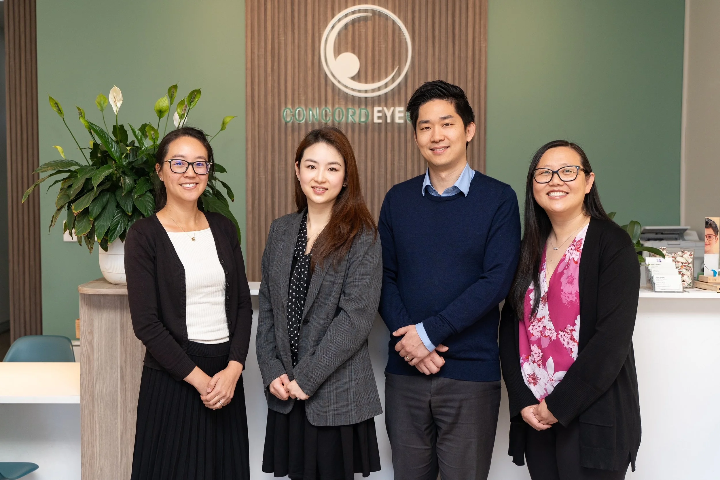 Four smiling employees standing in front of the Concord Eye logo at the reception area of an eye clinic.