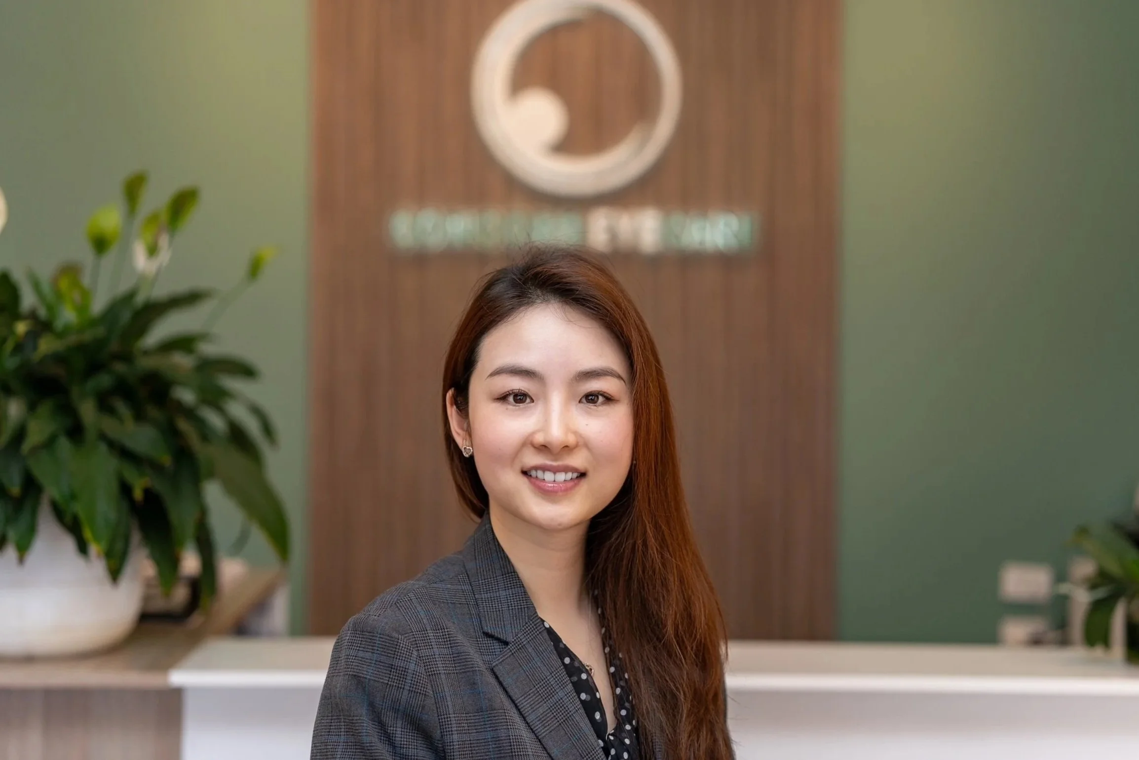 Optometrist Nikki Peng with long hair, wearing a blazer, smiling at the camera in an office reception area with a plant and a wooden wall with a logo in the background.