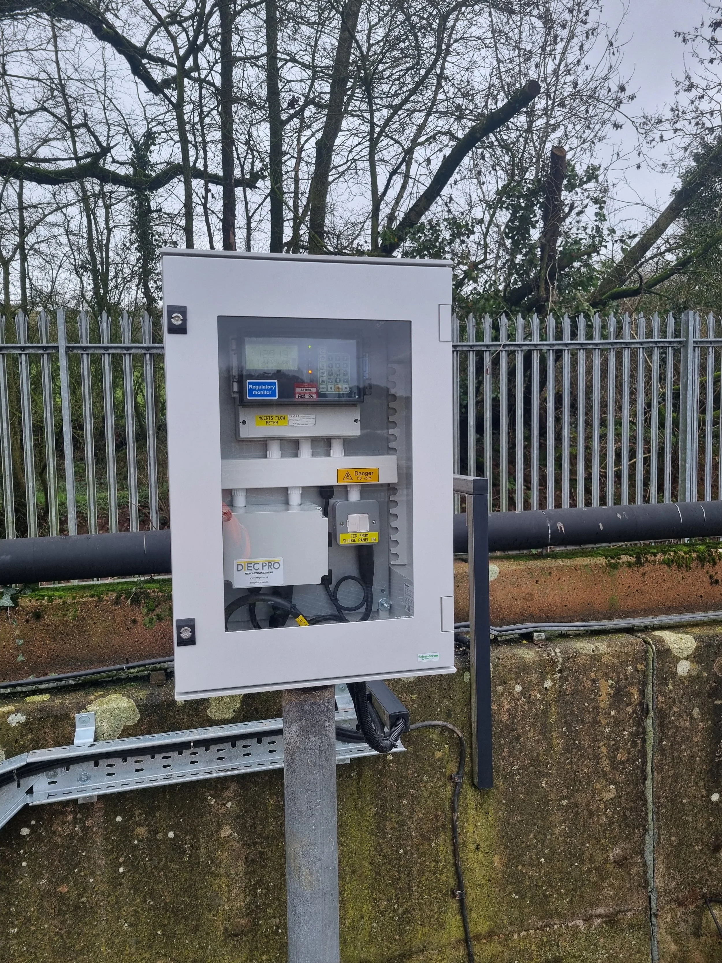 Electrical utility box mounted on a pole, with a fence and trees in the background.