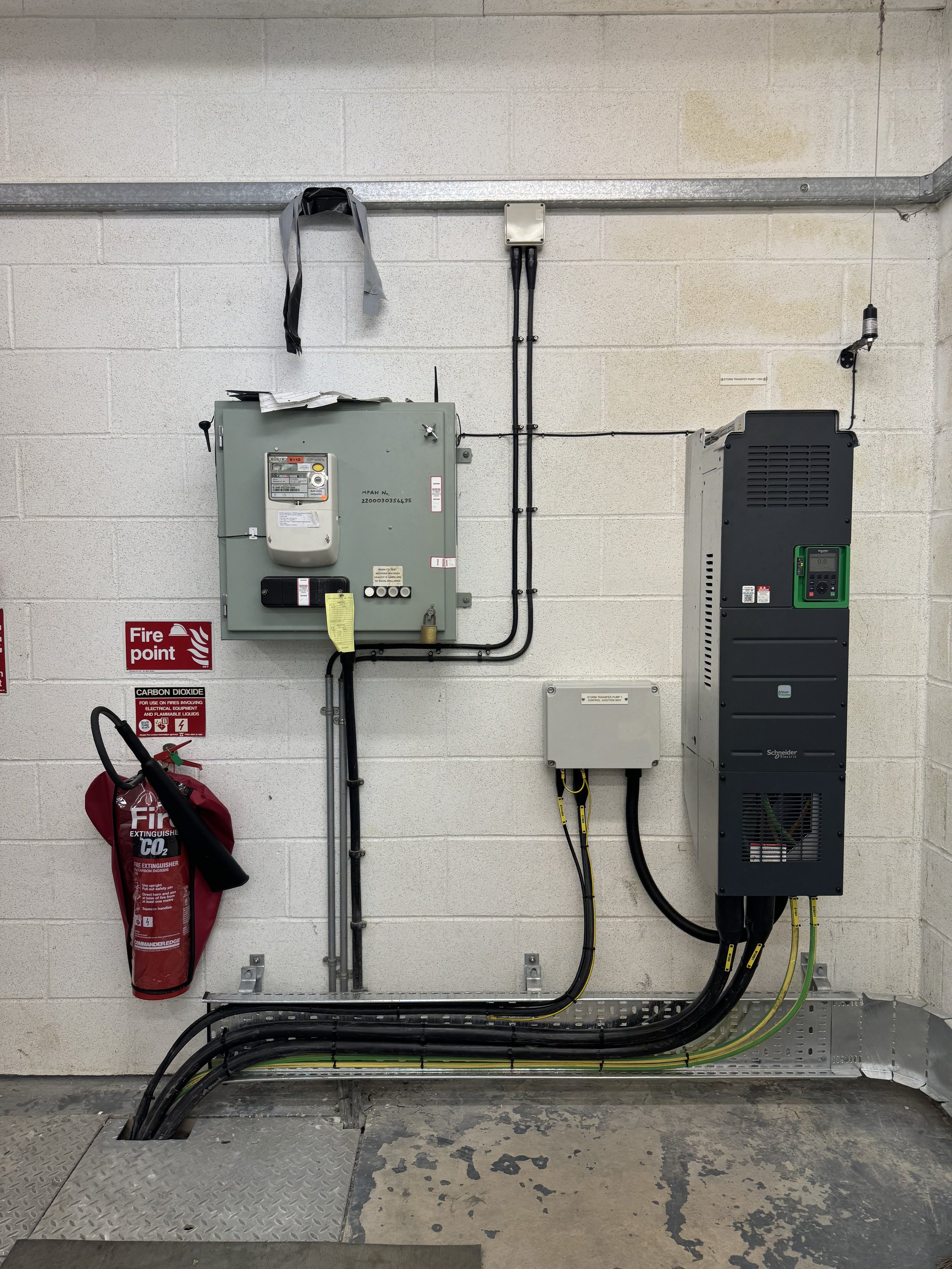 Electrical equipment and a fire extinguisher mounted on a beige brick wall with grey conduit pipes running from the top to the bottom.