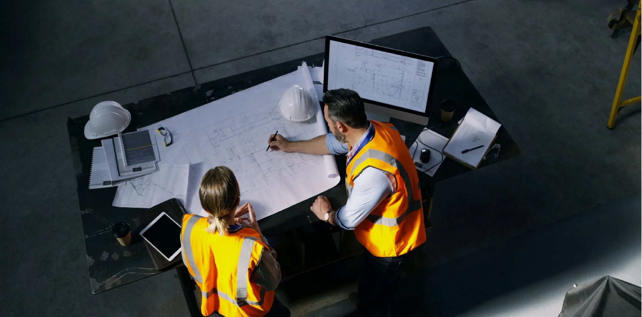 Two engineers in orange safety vests discussing blueprints at a table with technical drawings, laptops, and safety helmets in a construction or industrial setting.