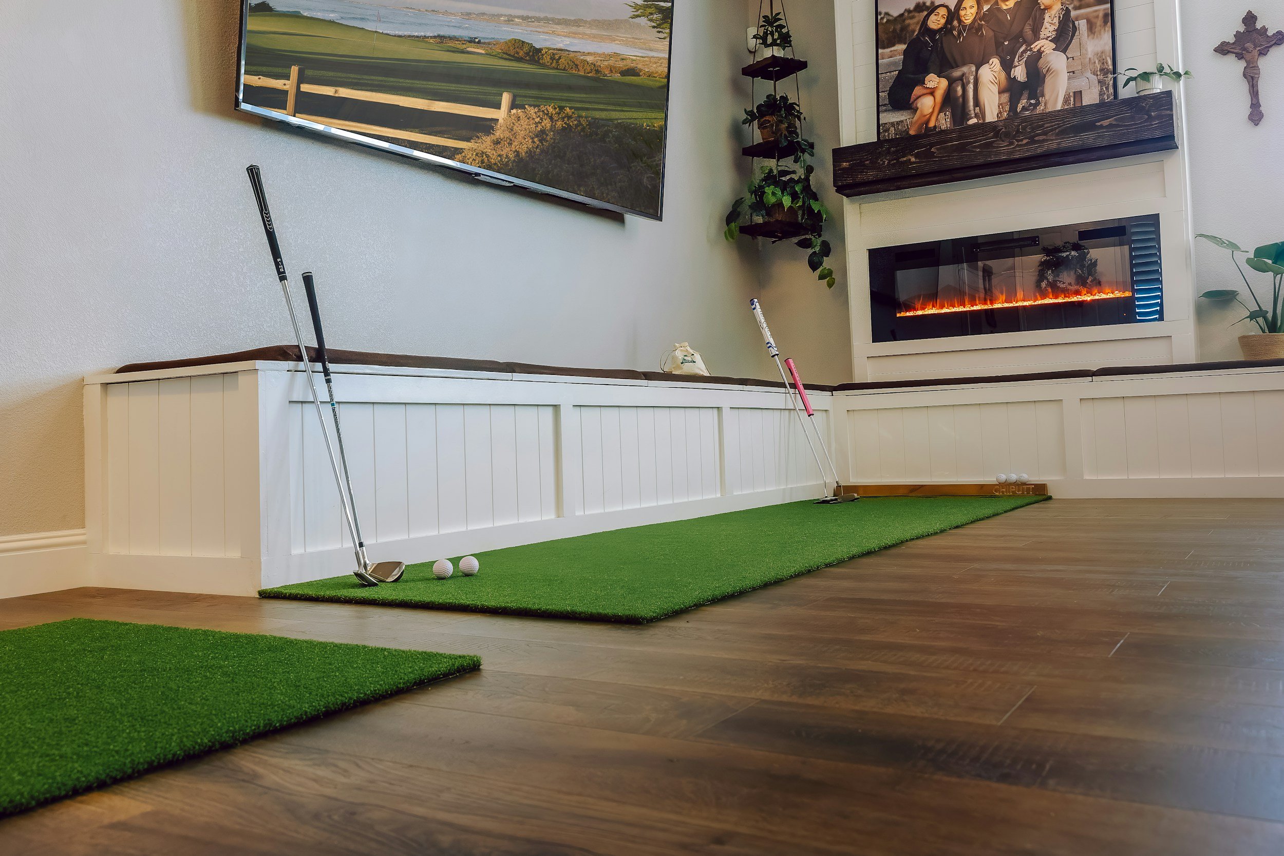 Indoor golf simulation room with artificial turf, golf clubs, golf balls, and a large flat-screen display showing a golf course. There is an electric fireplace below a framed photo of a group of friends, with decorations including plants and a religious cross on the wall.