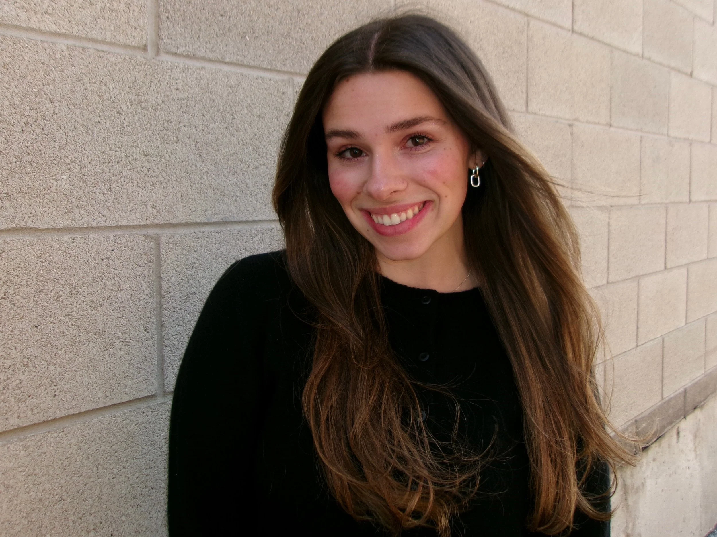 A young woman with long brown hair, wearing a black top and earrings, smiling while standing against a light-colored brick wall.