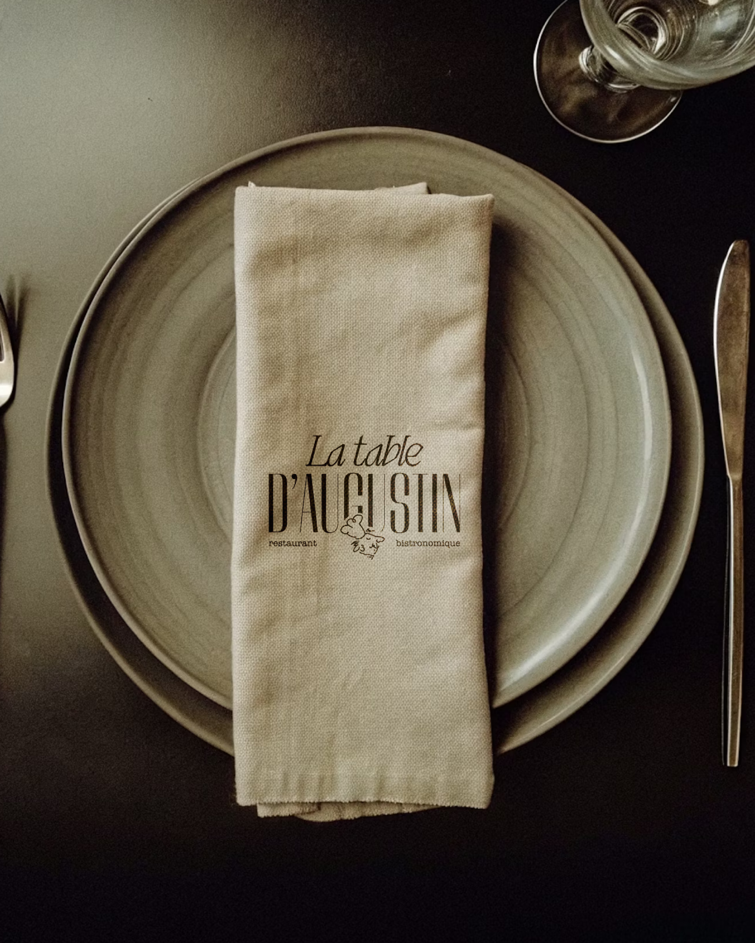 Une table de restaurant avec une grande assiette, un verre d'eau, un couteau et une serviette en tissu avec l'inscription 'La table D'AIG SIN', restaurant bistronomique.
