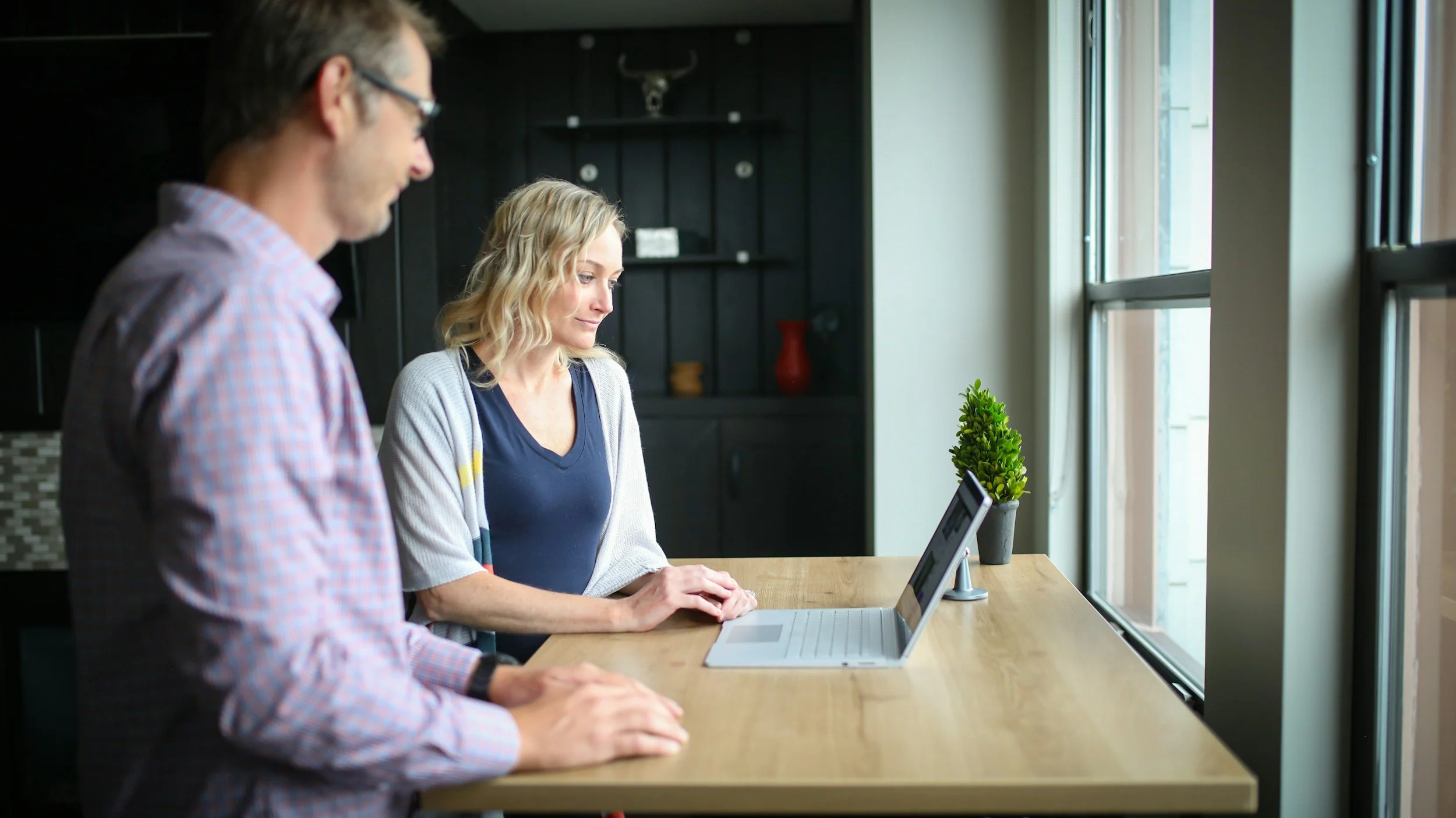 A woman and a man at a kitchen counter looking at a tablet, with a plant on the counter and a window beside them.