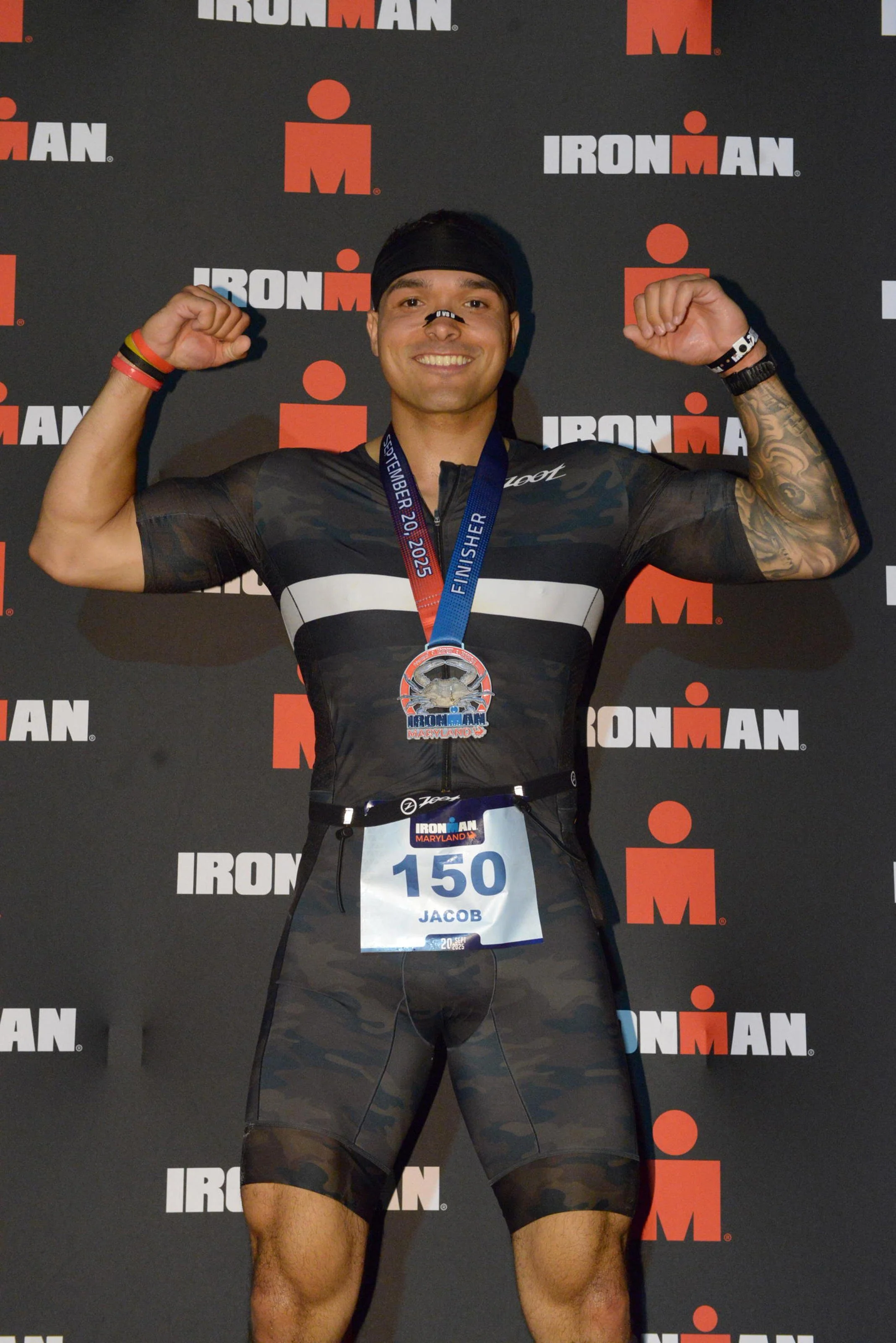 Male athlete with a medal around his neck, flexing his biceps, standing in front of an Ironman backdrop, wearing a black sports outfit and a race bib numbered 150 with the name Jacob.