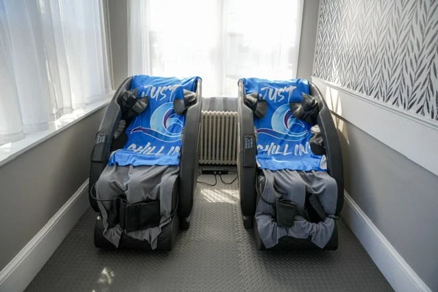 Two black baby car seats with blue Nike blankets draped over them, placed side by side in a bright room with white walls, window curtains, and a radiator between the seats.
