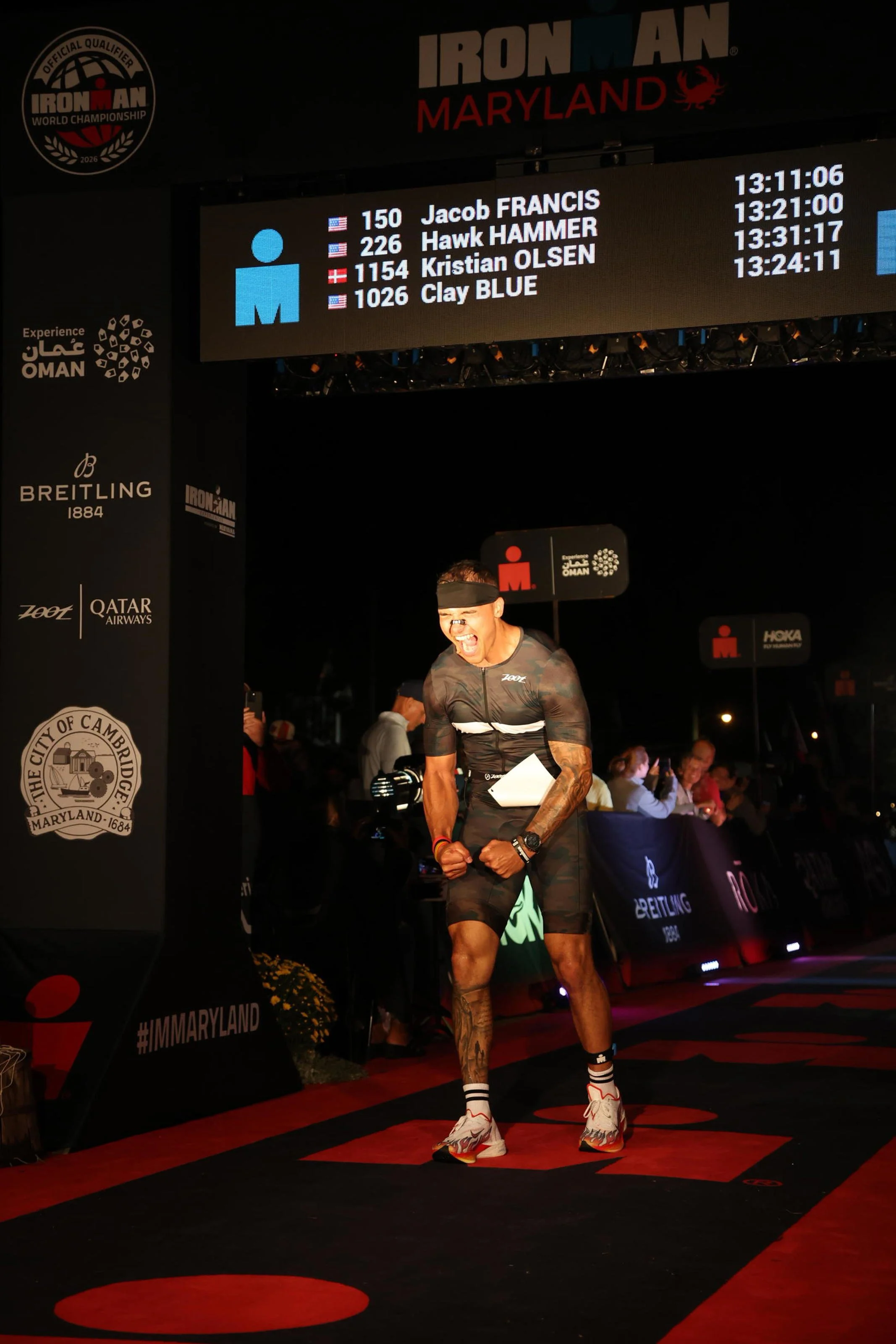 A male athlete celebrating as he crosses the finish line at the Ironman Maryland race, with a digital scoreboard above displaying his name and split times.