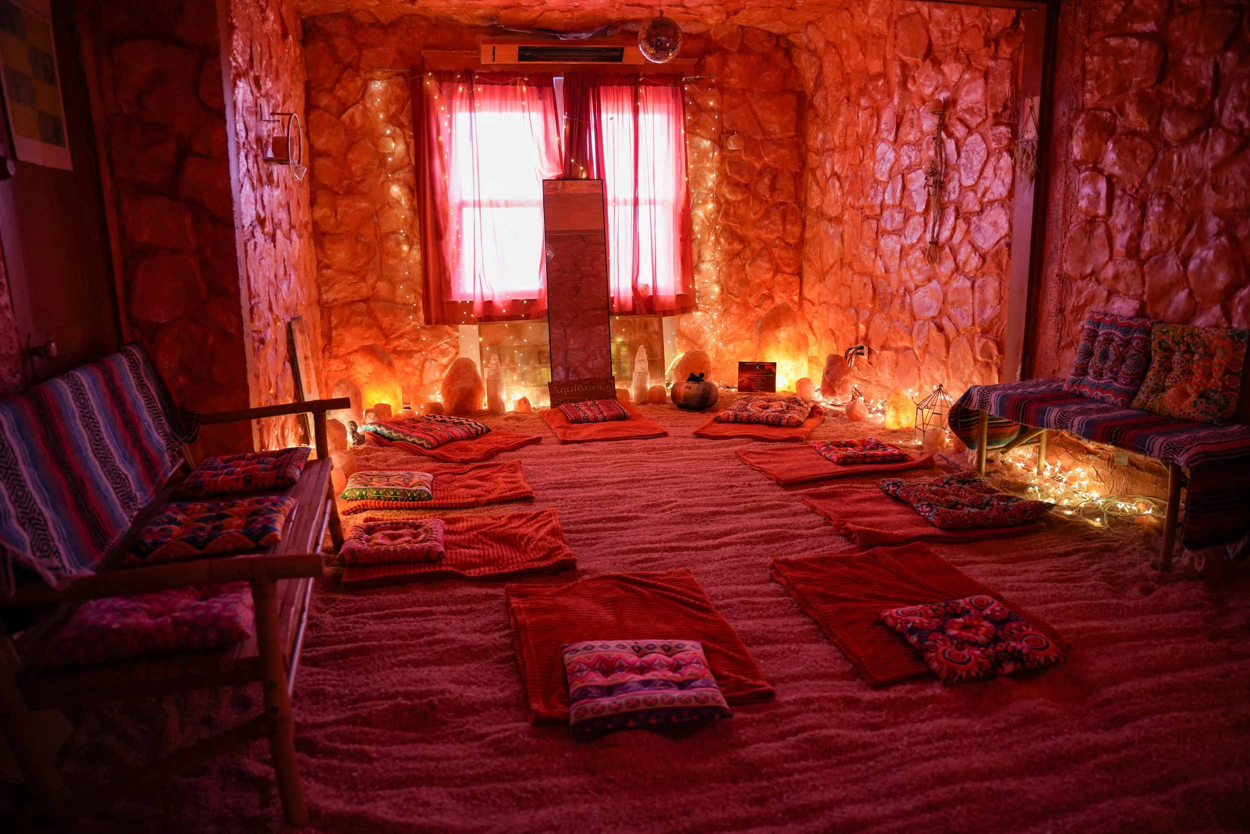 A warmly lit room with pink curtains, salt lamps, fairy lights, Himalayan salt decor, sandstone walls, and cushions arranged on a bed and benches. The room has a cozy and relaxing ambiance, with a circular seating arrangement, soft lighting, and earthy tones.