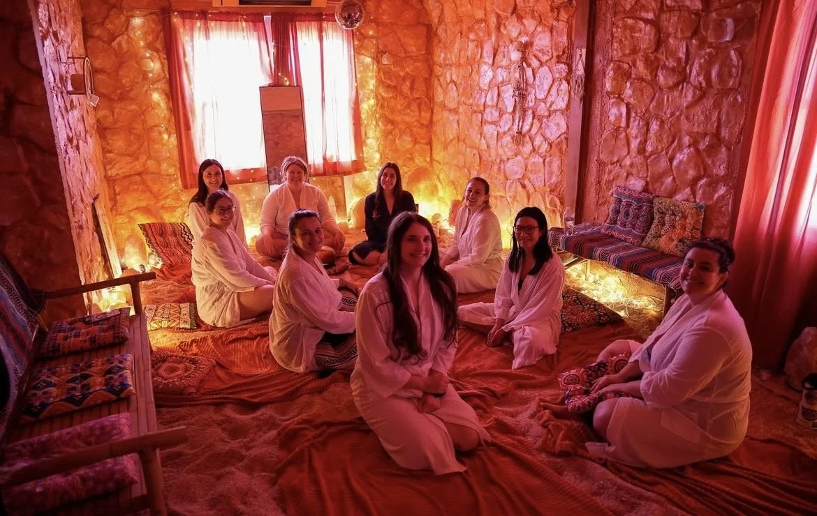 Ten women sitting on a cozy bed in a room with warm orange lighting, stone walls, and red curtains, some wearing white robes and some in dark clothes, smiling at the camera, surrounded by cushions and decorative pillows.