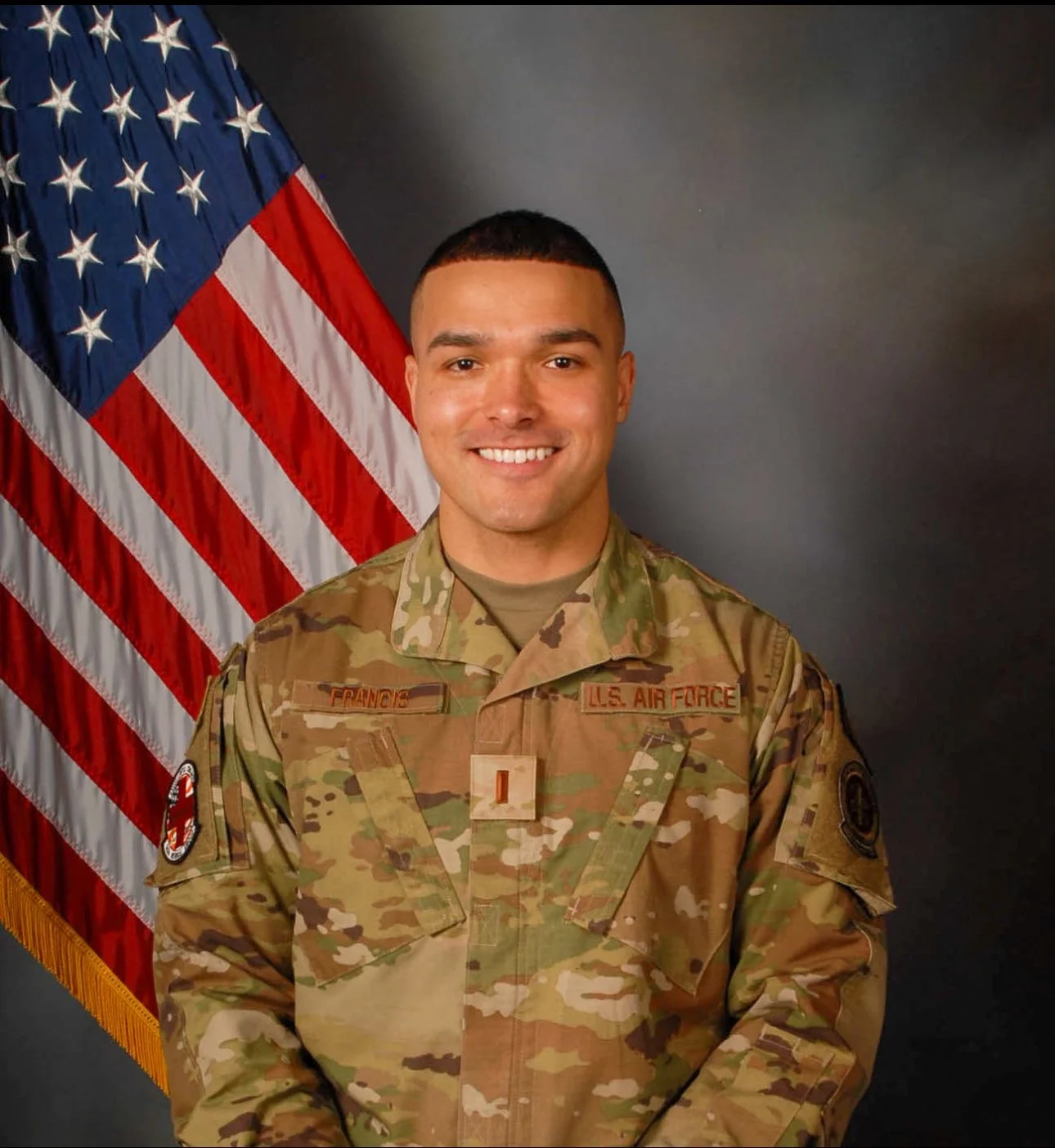 A smiling man in U.S. military uniform standing in front of an American flag.