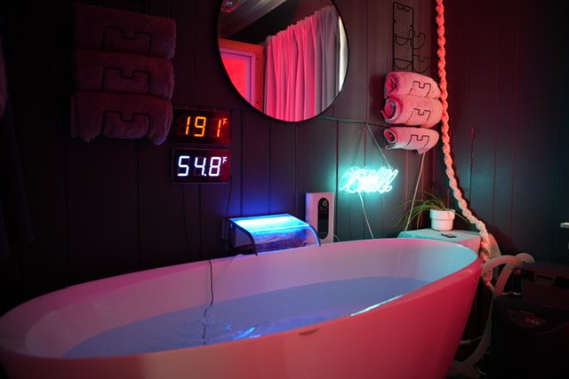 A cozy bathroom with a modern bathtub filled with water, dark wooden wall panels, a round mirror, and a neon sign. Digital displays show the temperature as 19°F and 54.8°F. Towels are rolled and hanging, with a small plant and a hairdryer nearby, illuminated by colorful lighting.
