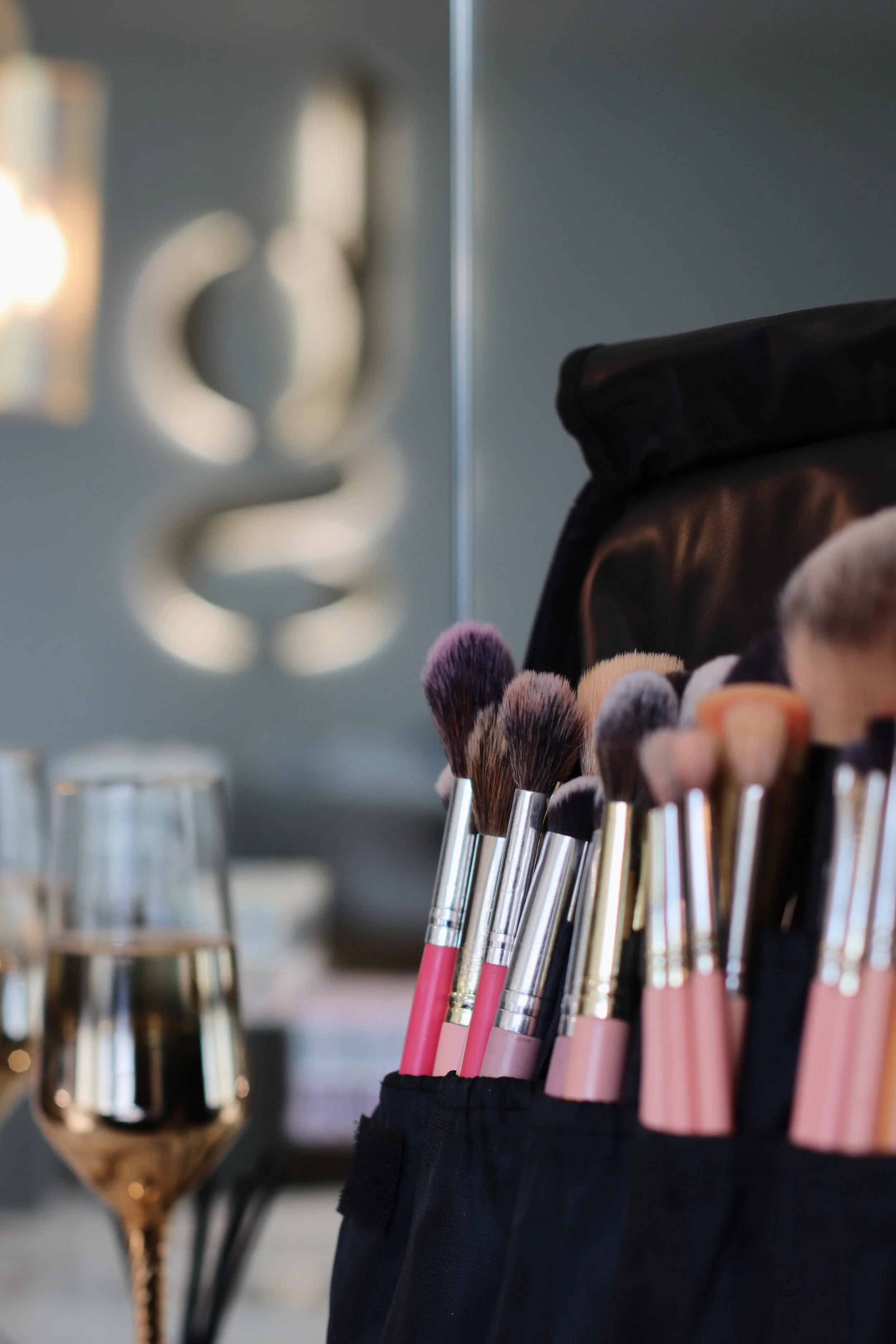 A collection of makeup brushes in a black pouch with two glasses of white wine in the foreground and a blurred wall decoration in the background.
