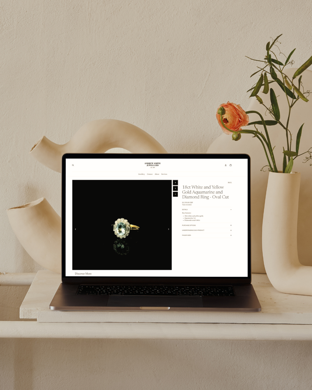 Open laptop on a white shelf displaying a jewelry website featuring a diamond ring, with a beige wall, abstract sculpture, and a white vase with pink and green flowers in the background.