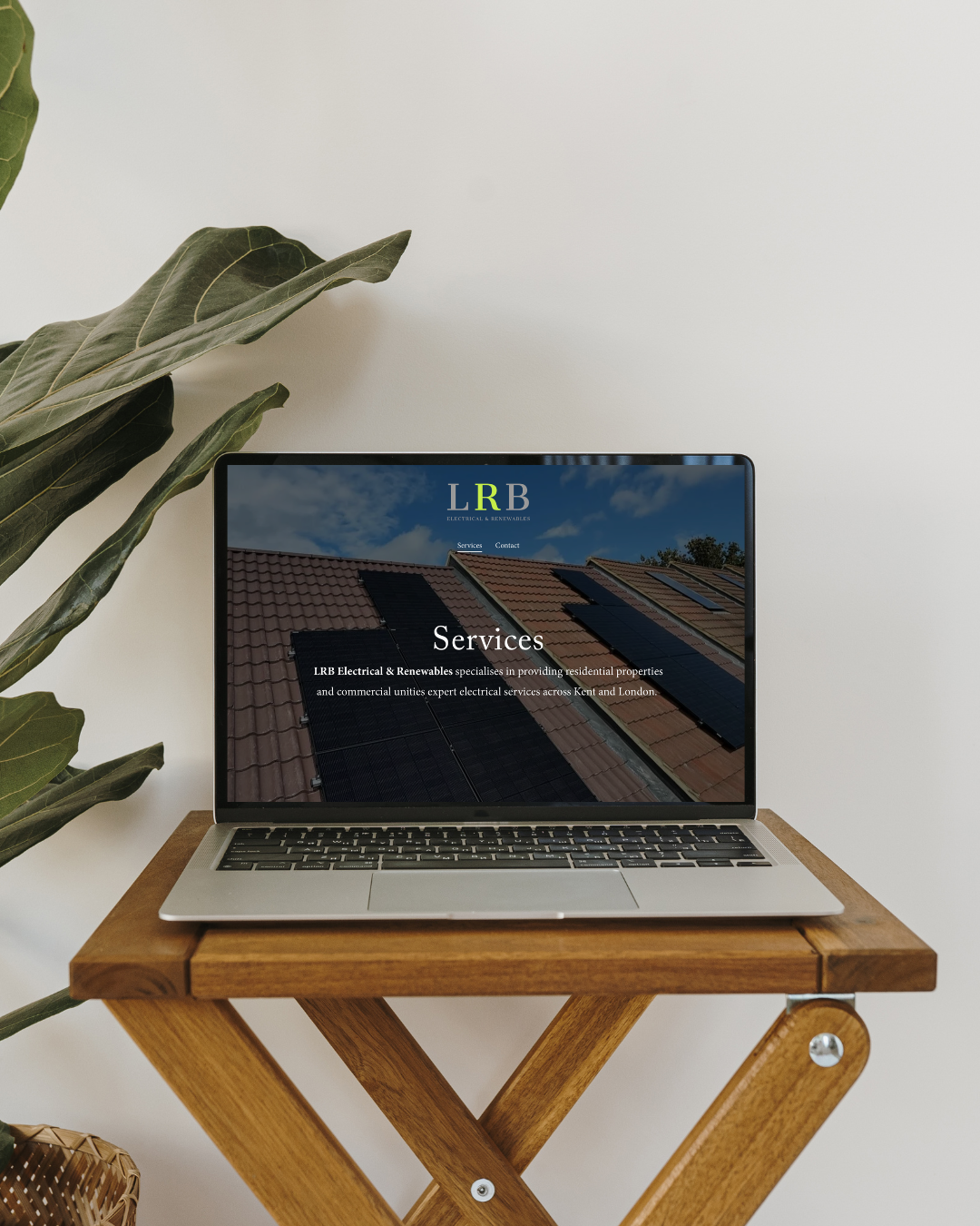 Laptop displaying a website for LRB Electrical & Renewables, placed on a wooden table with a large leafy plant nearby.