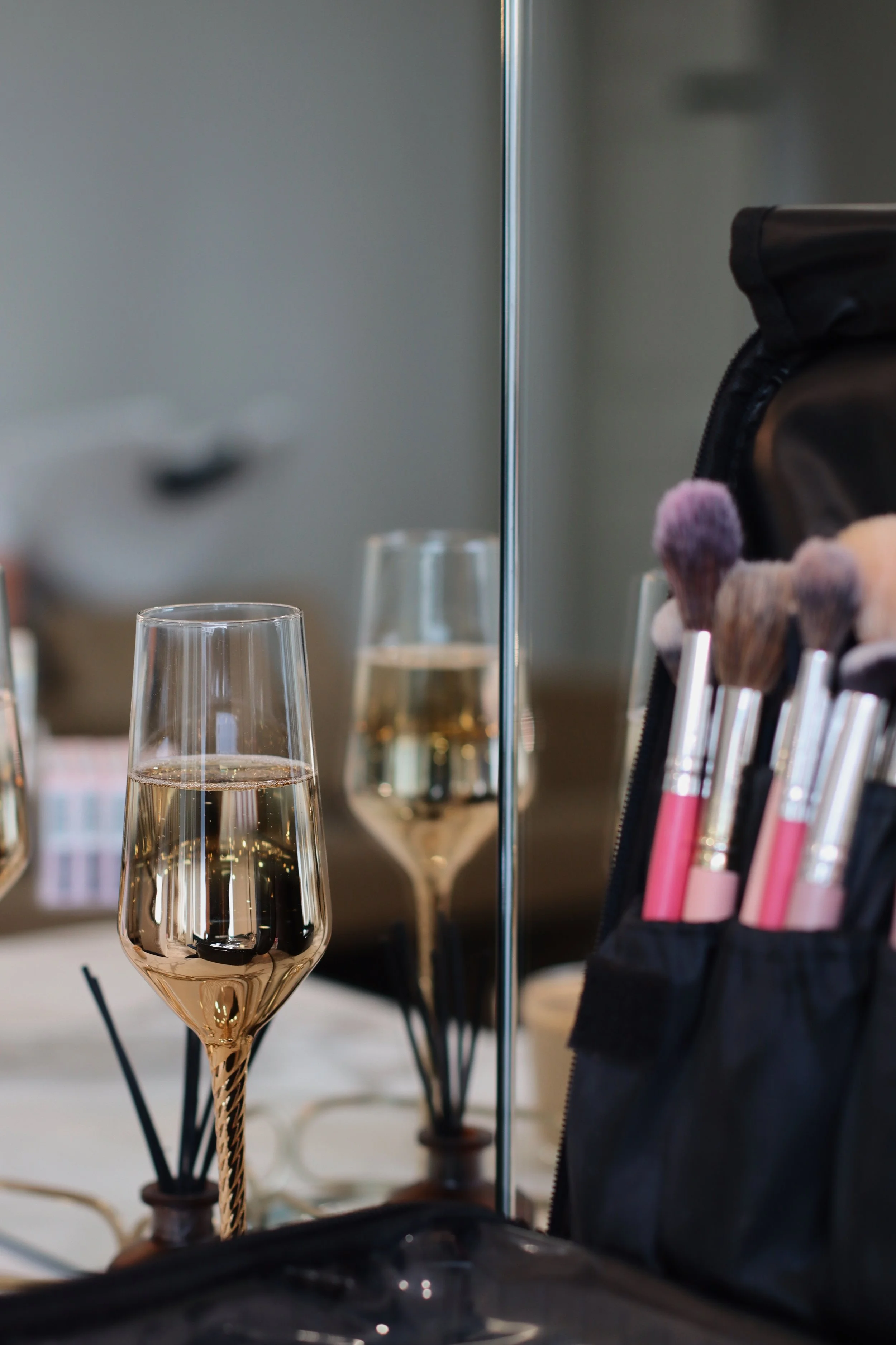 A mirror reflecting a champagne flute filled with a bubbly beverage, makeup brushes in a case, and a blurred background.