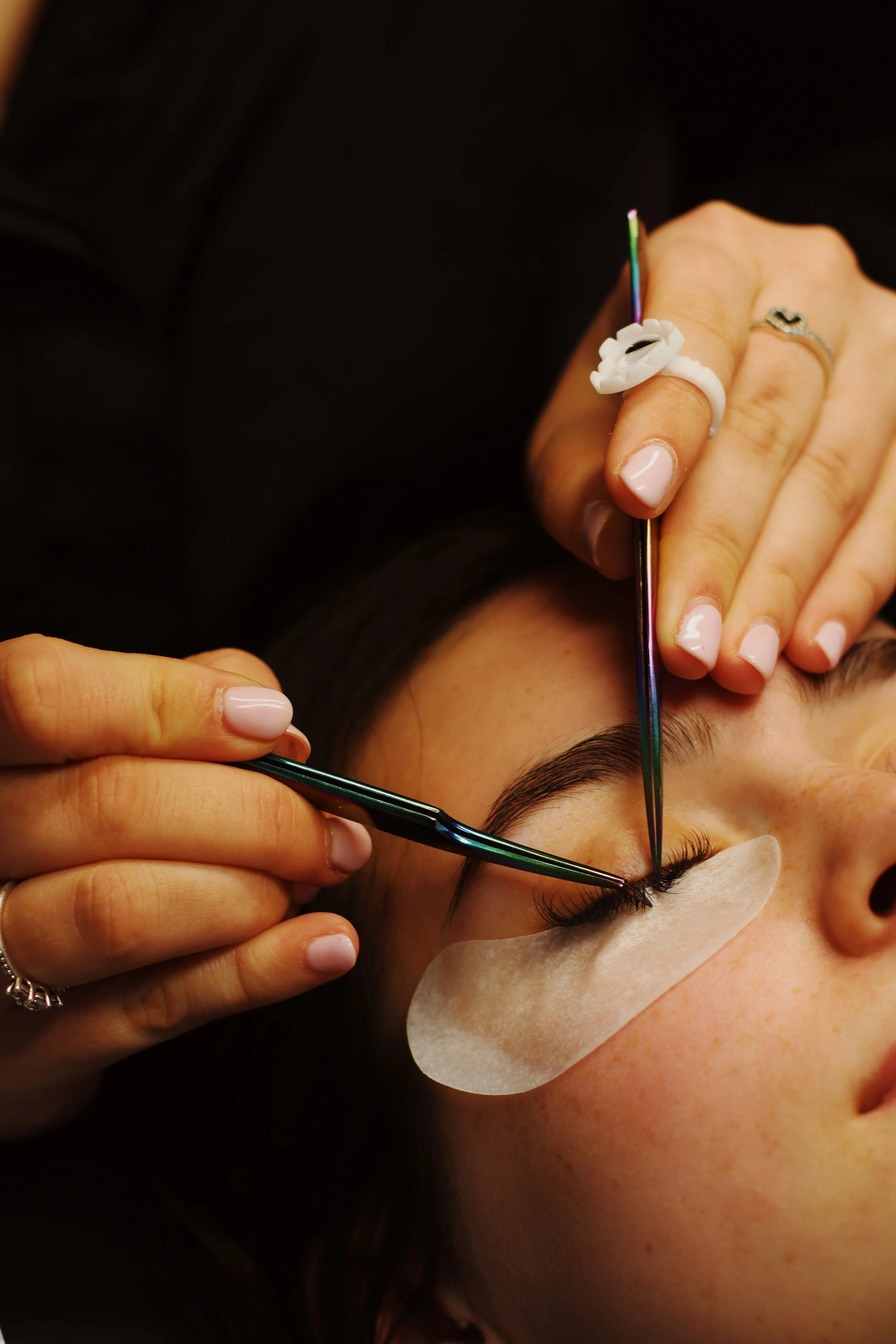 Close-up of a woman receiving eyelash extension treatment, with a technician applying false eyelashes with tweezers, and wearing a protective eye patch under her eye.