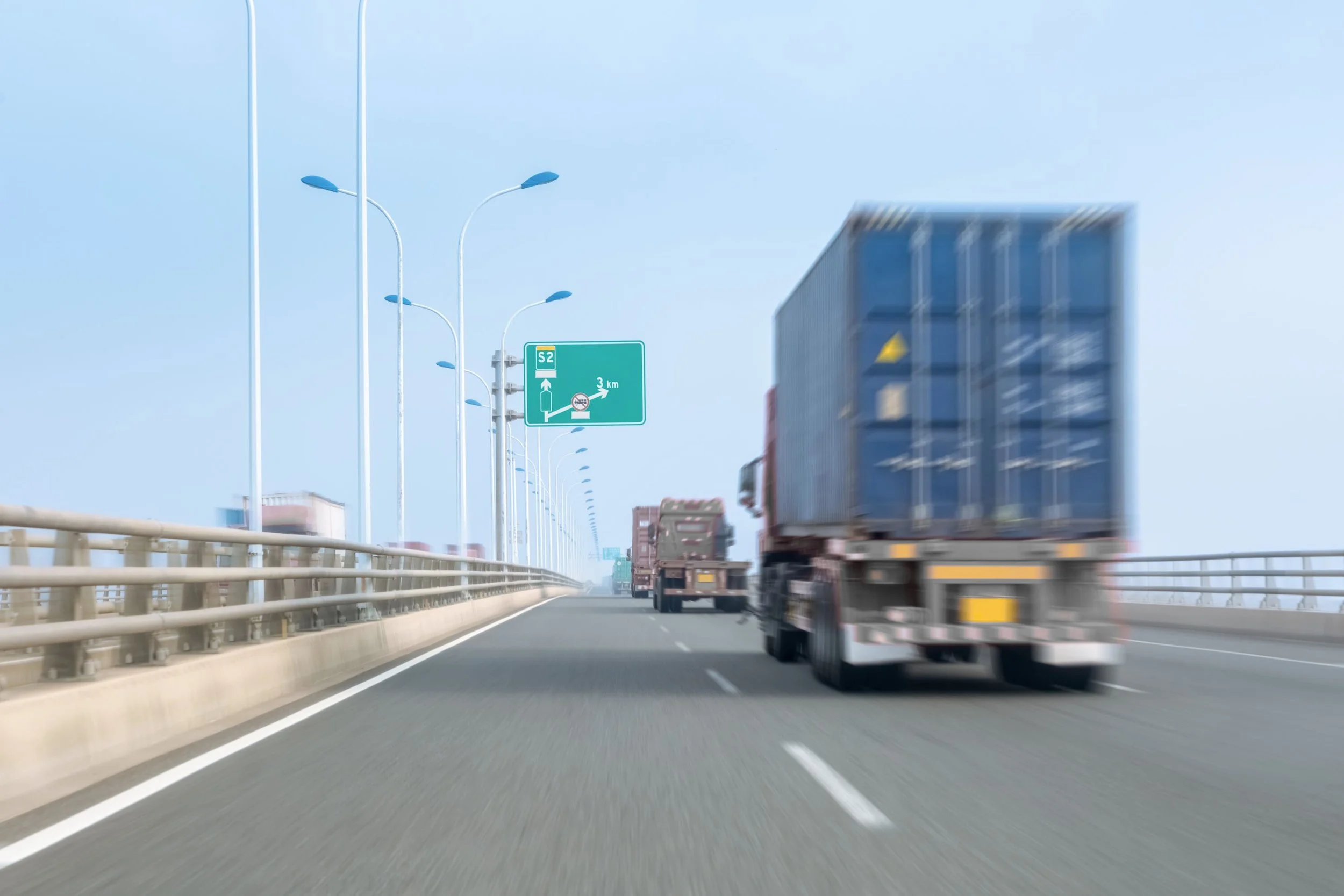 Auf einer Autobahn fahren mehrere LKWs in Fahrtrichtung, mit einer grünen Verkehrstafel und Straßenlaternen im Hintergrund.