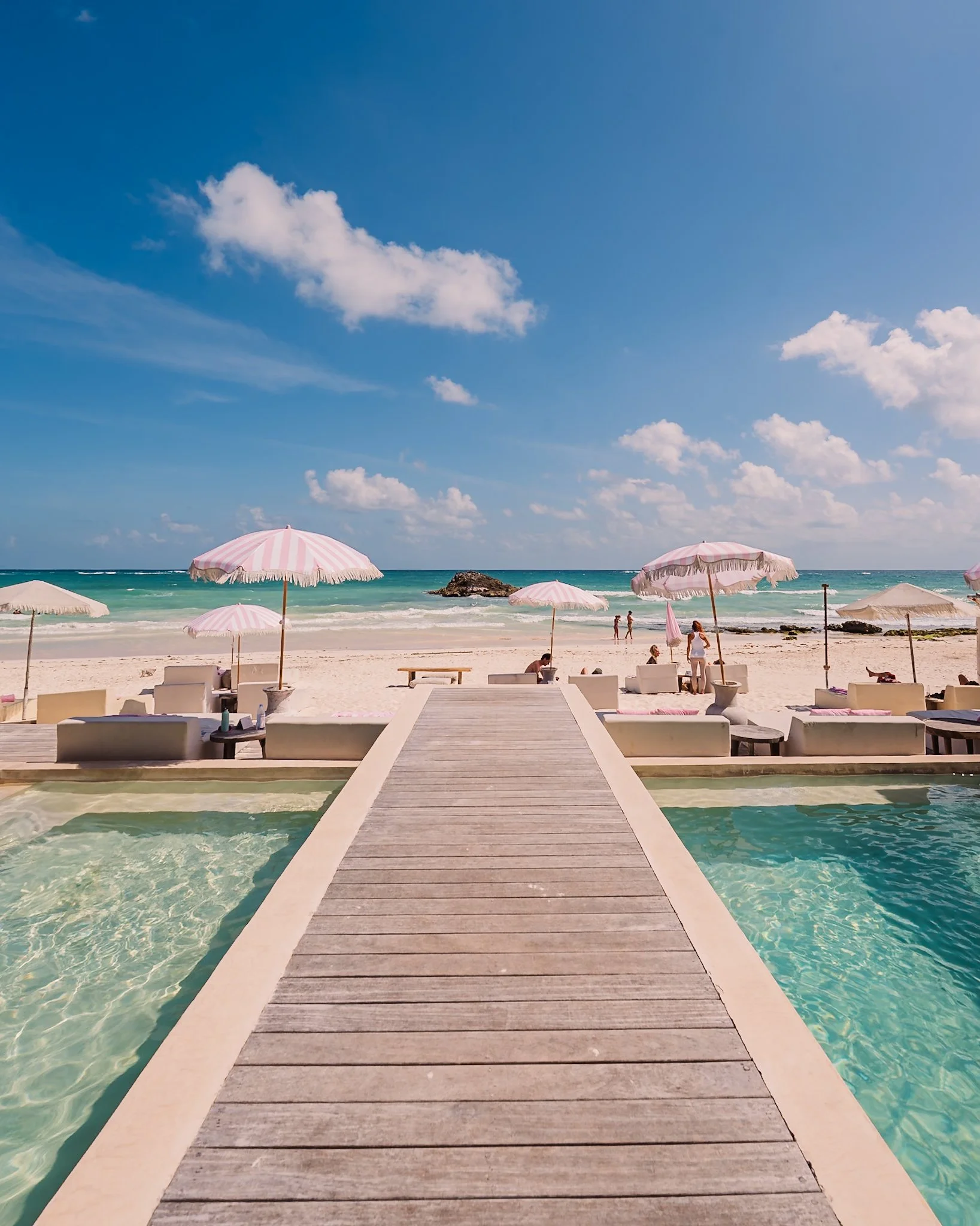 Beach scene with a wooden walkway leading to the sandy shore, surrounded by pink and white umbrellas, lounge chairs, and people enjoying the sunny day by the ocean under a bright blue sky with scattered clouds.