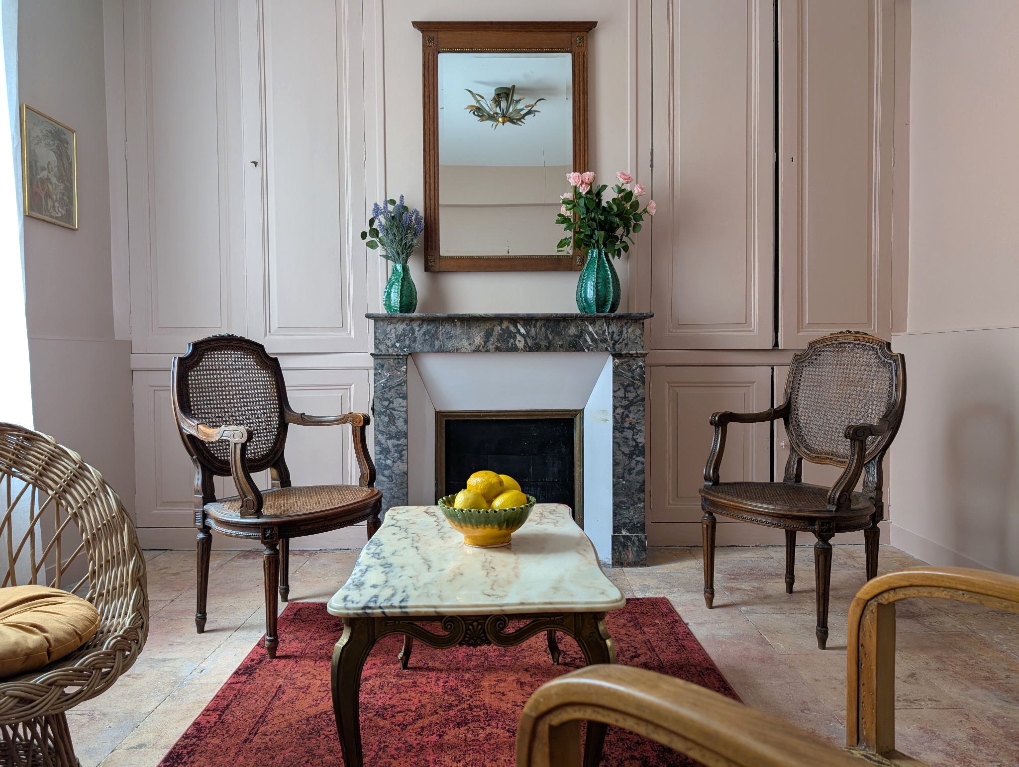 Room with a marble fireplace, wooden chairs, a red rug, a marble table, a bowl of lemons, vases with flowers, and a mirror