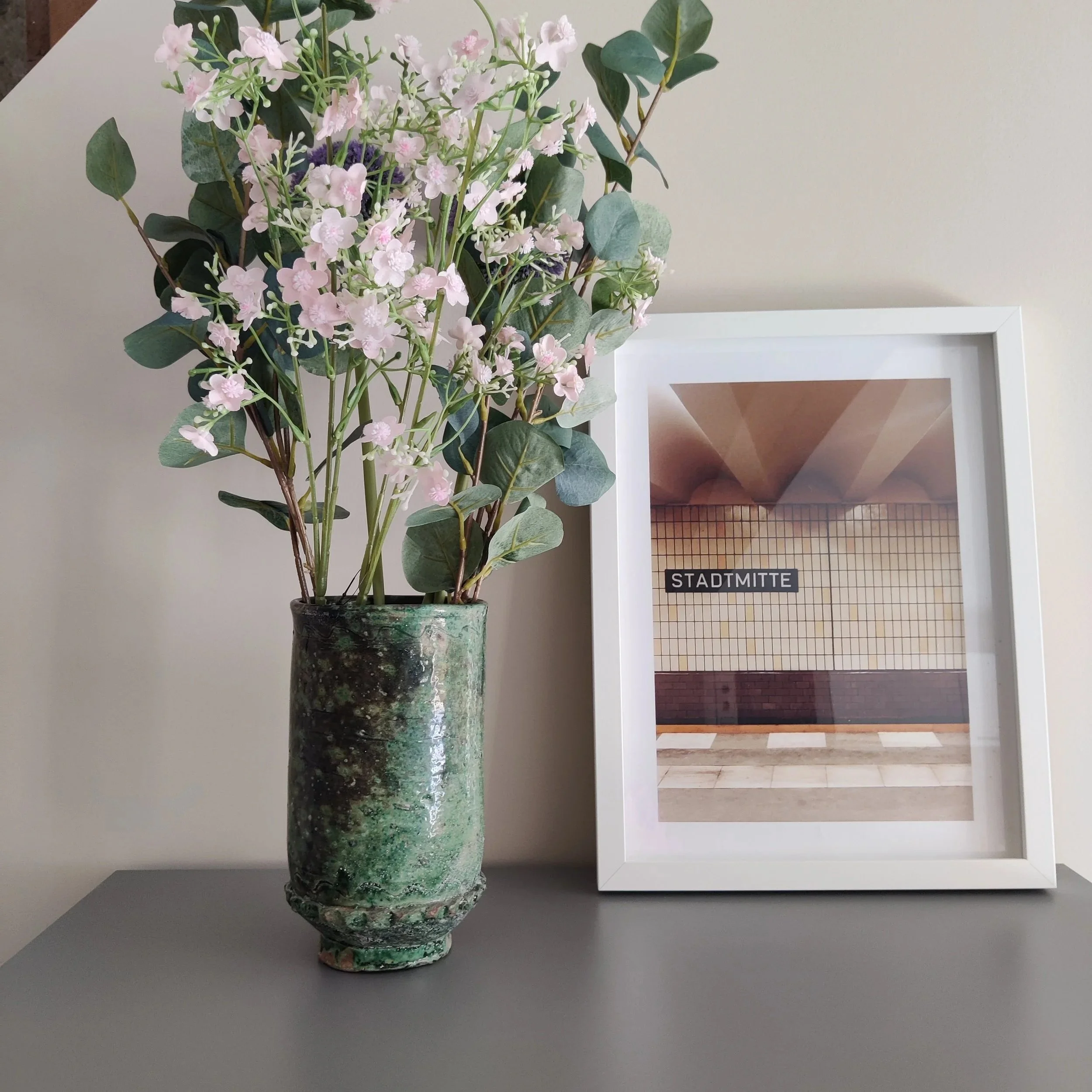 A green ceramic vase containing pink and green flowers, next to a white-framed photo of a subway station titled 'STADTMITTE'.