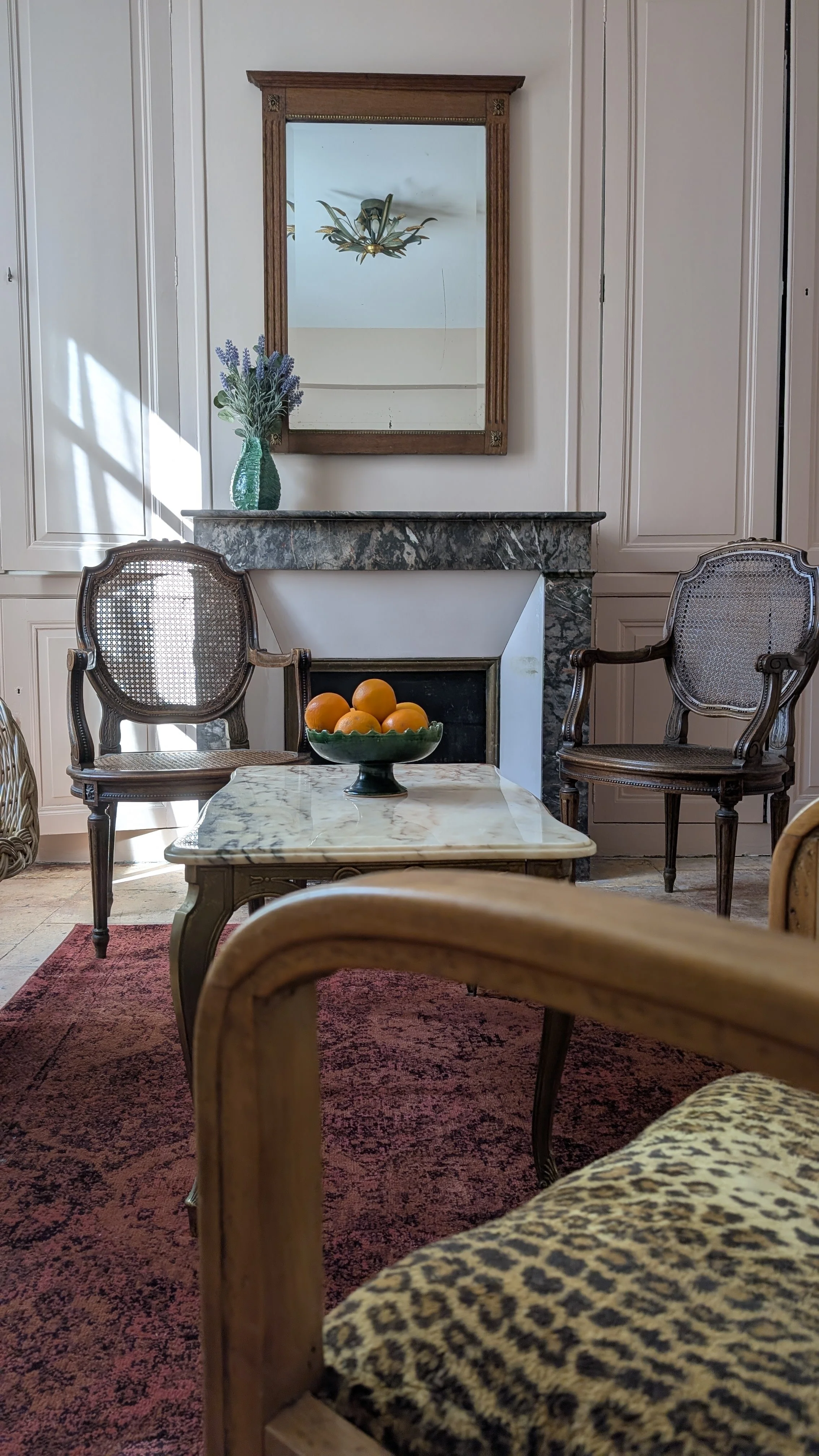 Elegant room, antique mirror, marble fireplace,  wooden armchairs with cane-backed seats, marble table with bowl of oranges, vase of purple flowers, dark red rug.