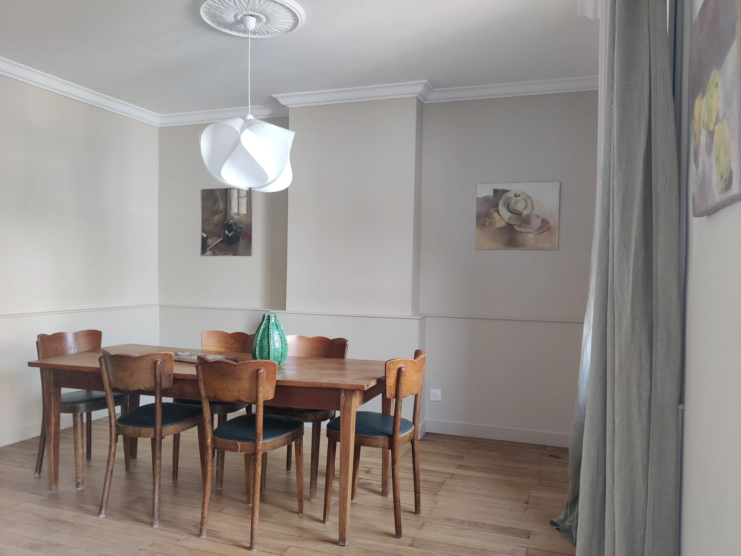 Dining room, wooden table, wooden chairs, vase in the center of the table, paintings on the wall, modern white ceiling light, hardwood floors