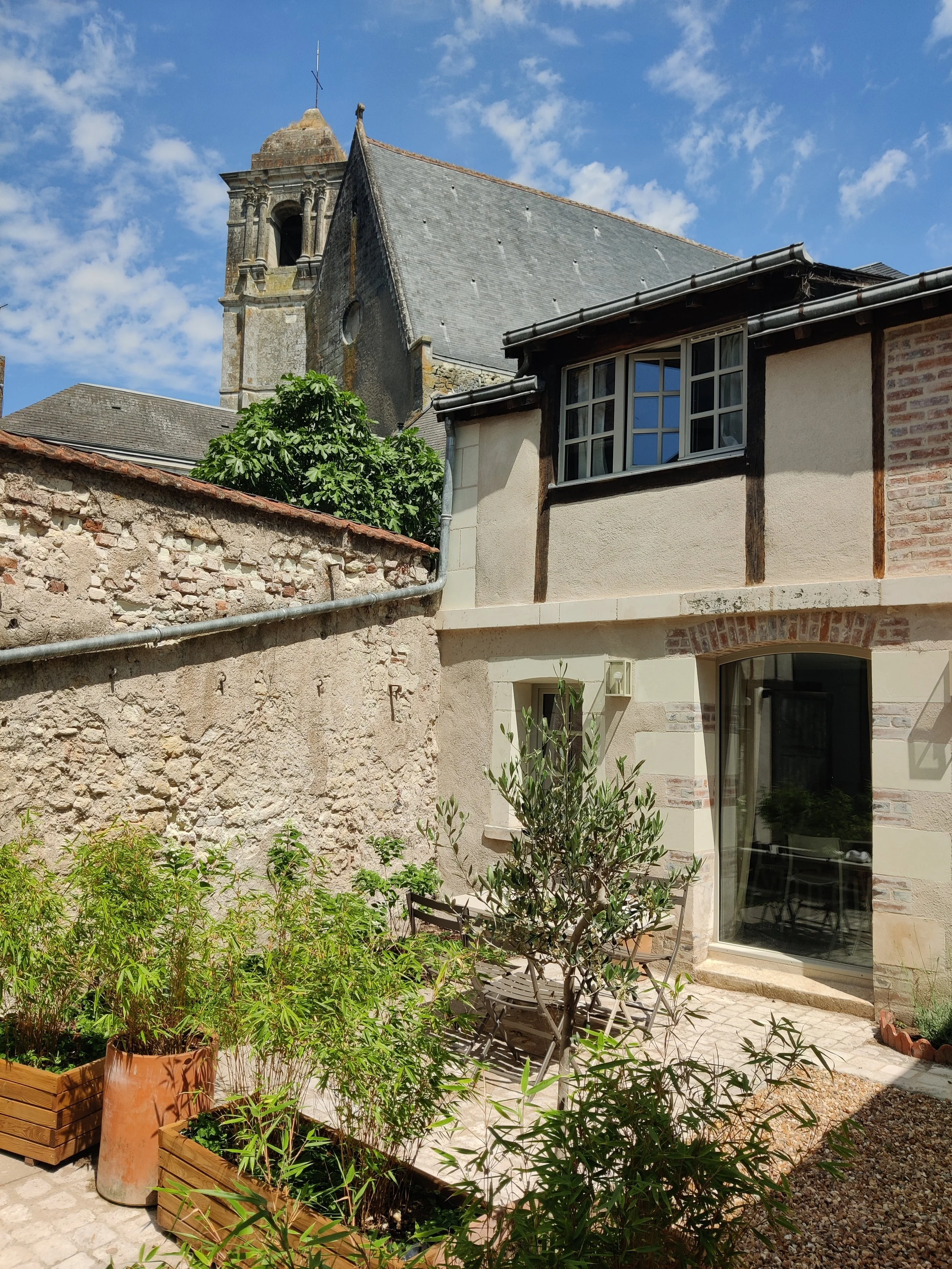 An old building with a stone facade and a slate roof, a large paned window, a small garden with potted plants, and an outdoor table. In the background, a stone church with a bell tower.