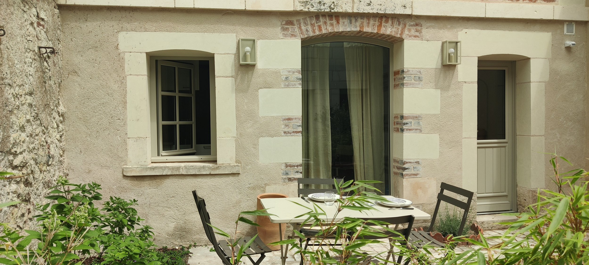 Photo of a stone house with an open window, a door, and a patio with a table and chairs, surrounded by greenery
