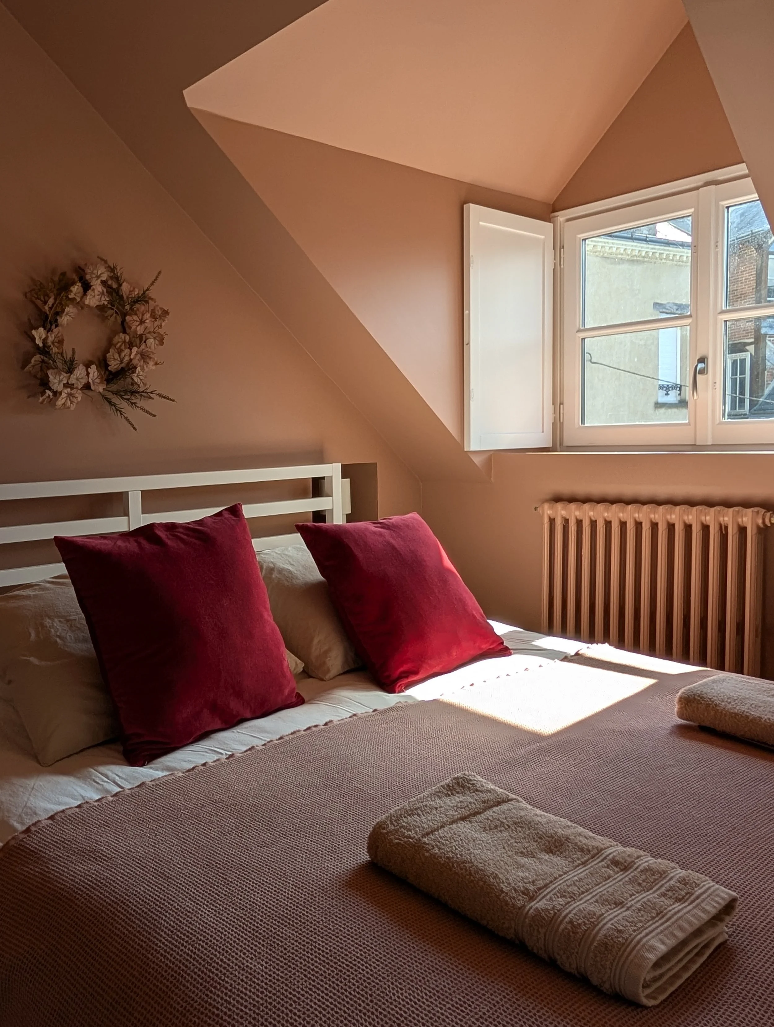 A bedroom with a bed, two red cushions, a window letting in light, a radiator underneath, and a wall decoration shaped like a wreath of flowers.
