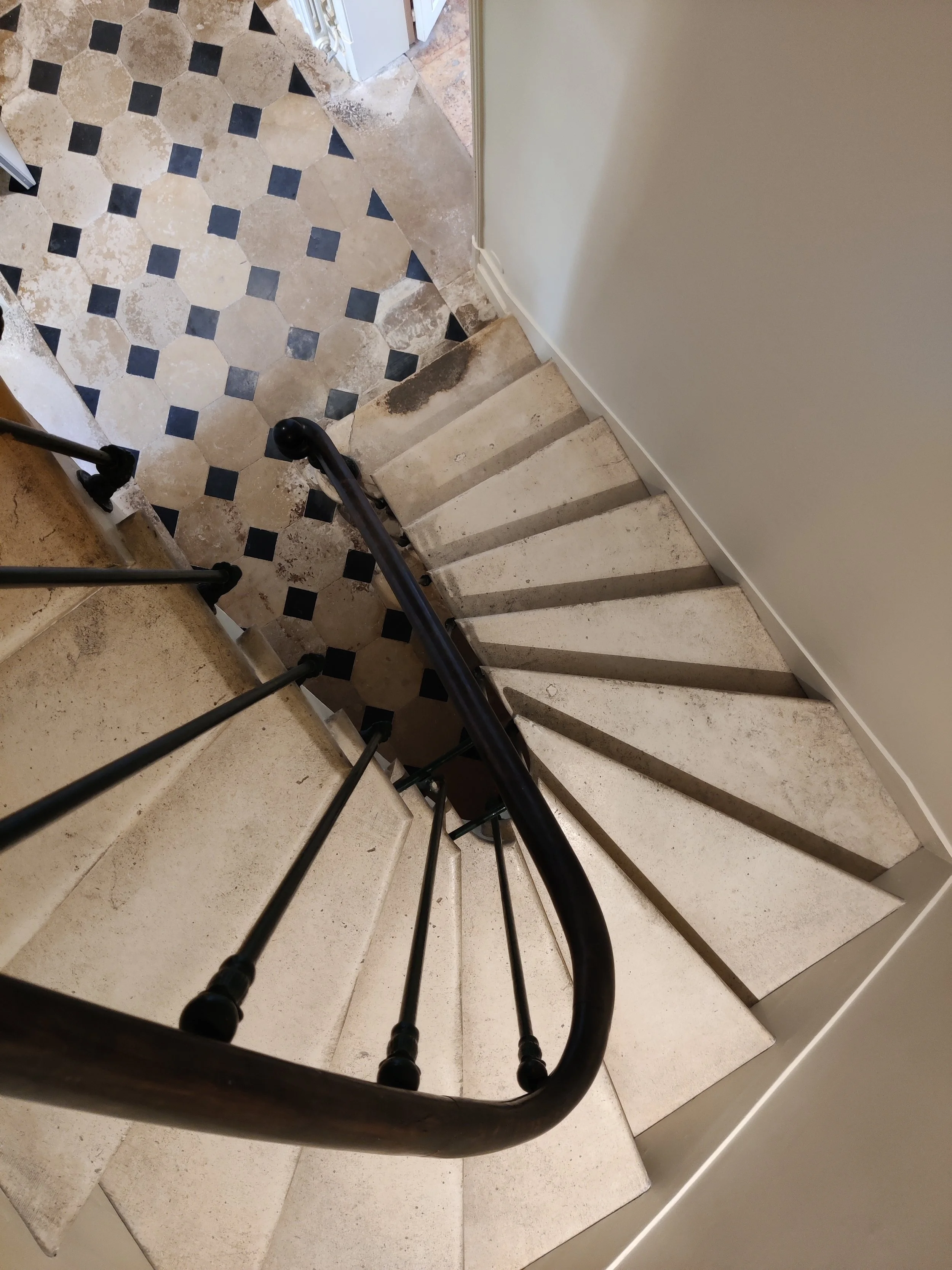 A stone spiral staircase with a black metal railing in a house, featuring beige and black tile flooring.